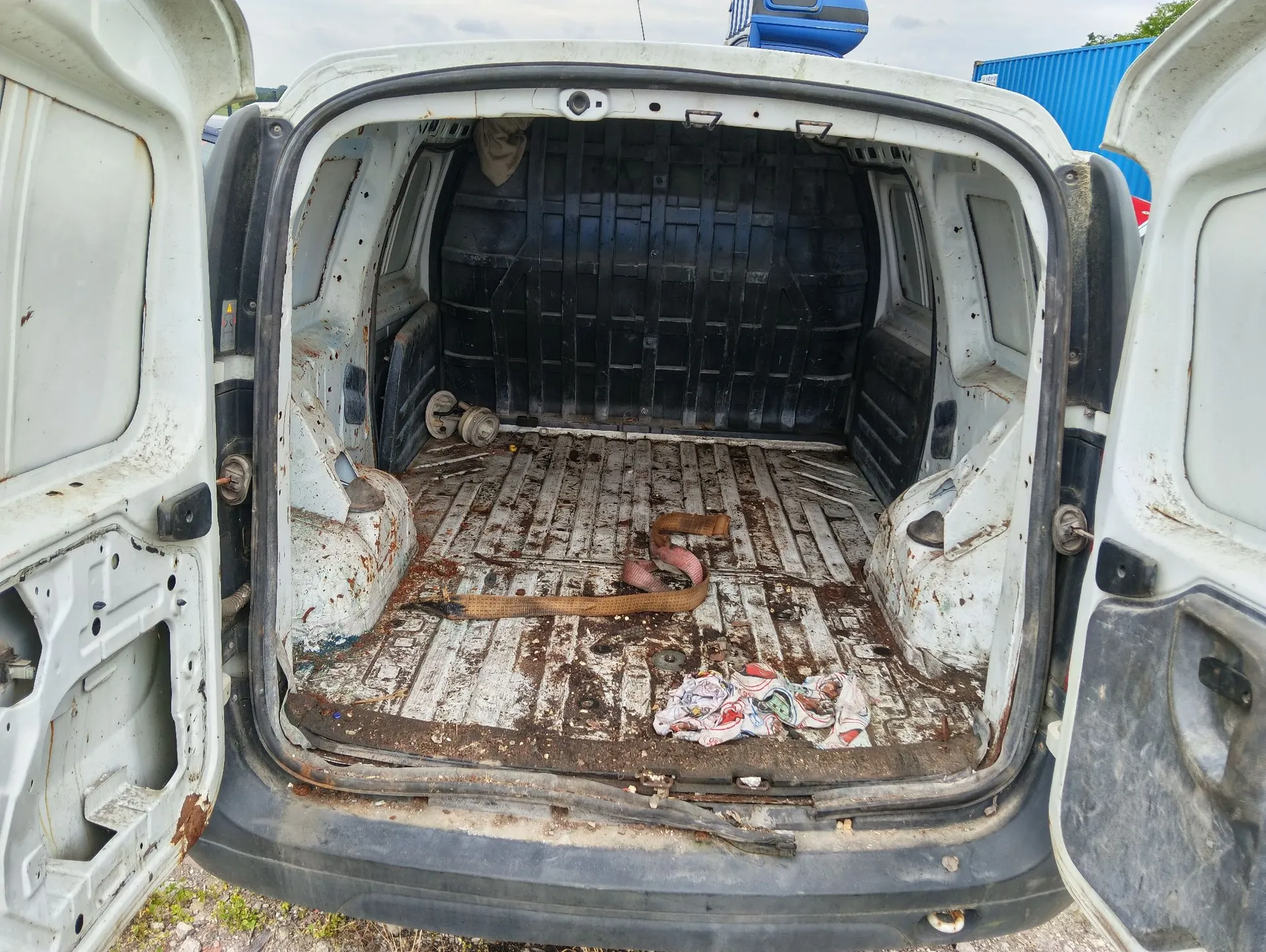 Widok z tyłu do wnętrza białego samochodu dostawczego z otwartymi tylnymi drzwiami. Przestrzeń ładunkowa jest brudna, z widocznymi śladami rdzy i zabrudzeniami na podłodze. Wewnątrz znajduje się zwinięty, brązowy wąż lub kabel oraz kawałek brudnej, jasnej szmatki. Ścianki boczne i tylna przegroda są wykonane z ciemnego tworzywa sztucznego.