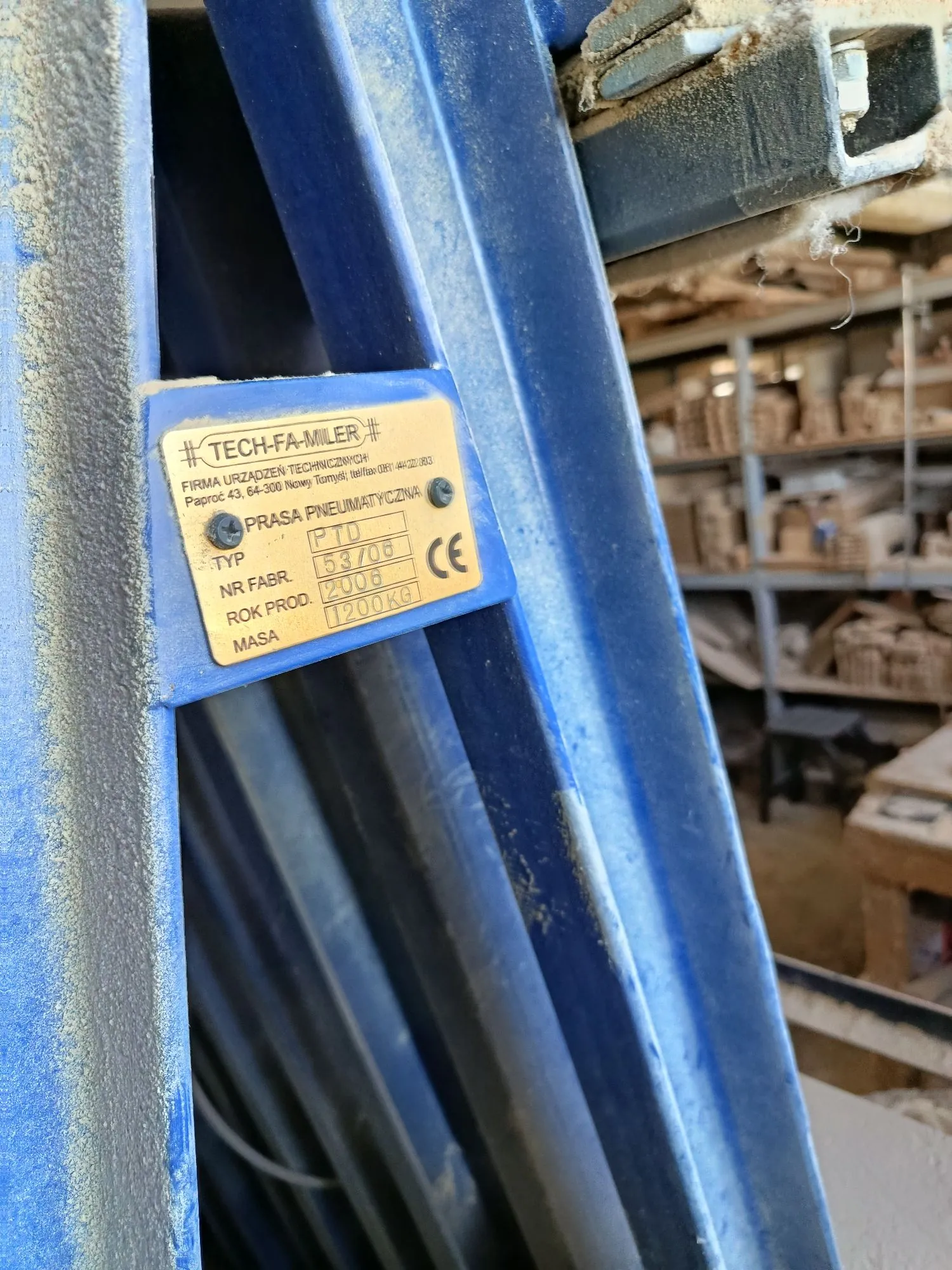 Zbliżenie na niebieską, metalową konstrukcję maszyny, do której przykręcona jest srebrna tabliczka znamionowa. Na tabliczce znajduje się tekst: '# TECH-FA-MILER #', 'FIRMA URZĄDZEŃ TECHNICZNYCH', 'Paproć 43, 64-500 Nieciecz Tomyśl, tel/fax: 061 44 02 00 03', 'PRASA PNEUMATYCZNA', 'TYP PTD 53/08', 'NR FABR. 2008', 'ROK PROD. 2008', 'MASA 1200KG' oraz znak CE.
