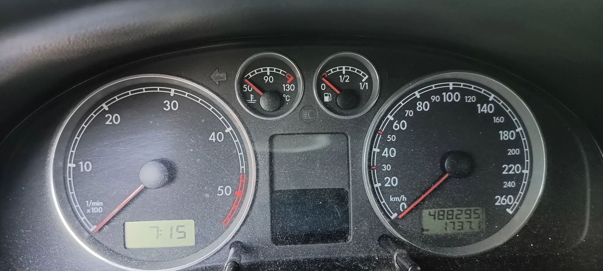 Zbliżenie na deskę rozdzielczą samochodu. Po lewej stronie znajduje się obrotomierz z cyframi 10, 20, 30, 40, 50 oraz oznaczeniem 1/min x 100 i cyfrowym zegarem wyświetlającym godzinę 7:15. W centrum znajdują się dwa mniejsze wskaźniki: temperatura cieczy chłodzącej (skala od 50 do 130 stopni Celsjusza) oraz poziom paliwa (skala od 0 do 1/1). Po prawej stronie znajduje się prędkościomierz z cyframi od 0 do 260 km/h oraz cyfrowym licznikiem przebiegu wyświetlającym wartość 488295. Pomiędzy wskaźnikami widoczne są kontrolki ostrzegawcze.