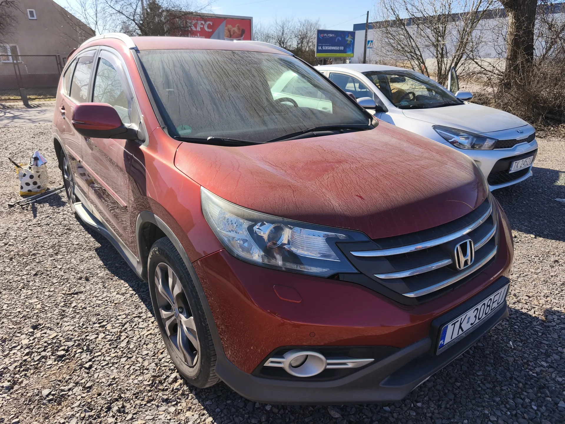 Na zdjęciu widoczny jest czerwony samochód osobowy marki Honda z tablicą rejestracyjną TK 308EU, zaparkowany na żwirowym placu. Za nim stoi biały samochód z tablicą TK 2452. W tle znajdują się budynki, w tym sklep KFC z czerwonym szyldem oraz niebieski billboard. Po lewej stronie obrazu leży torba w kropki i narzędzia. Drzewa bez liści oraz jasne niebo wskazują na pogodny dzień.
