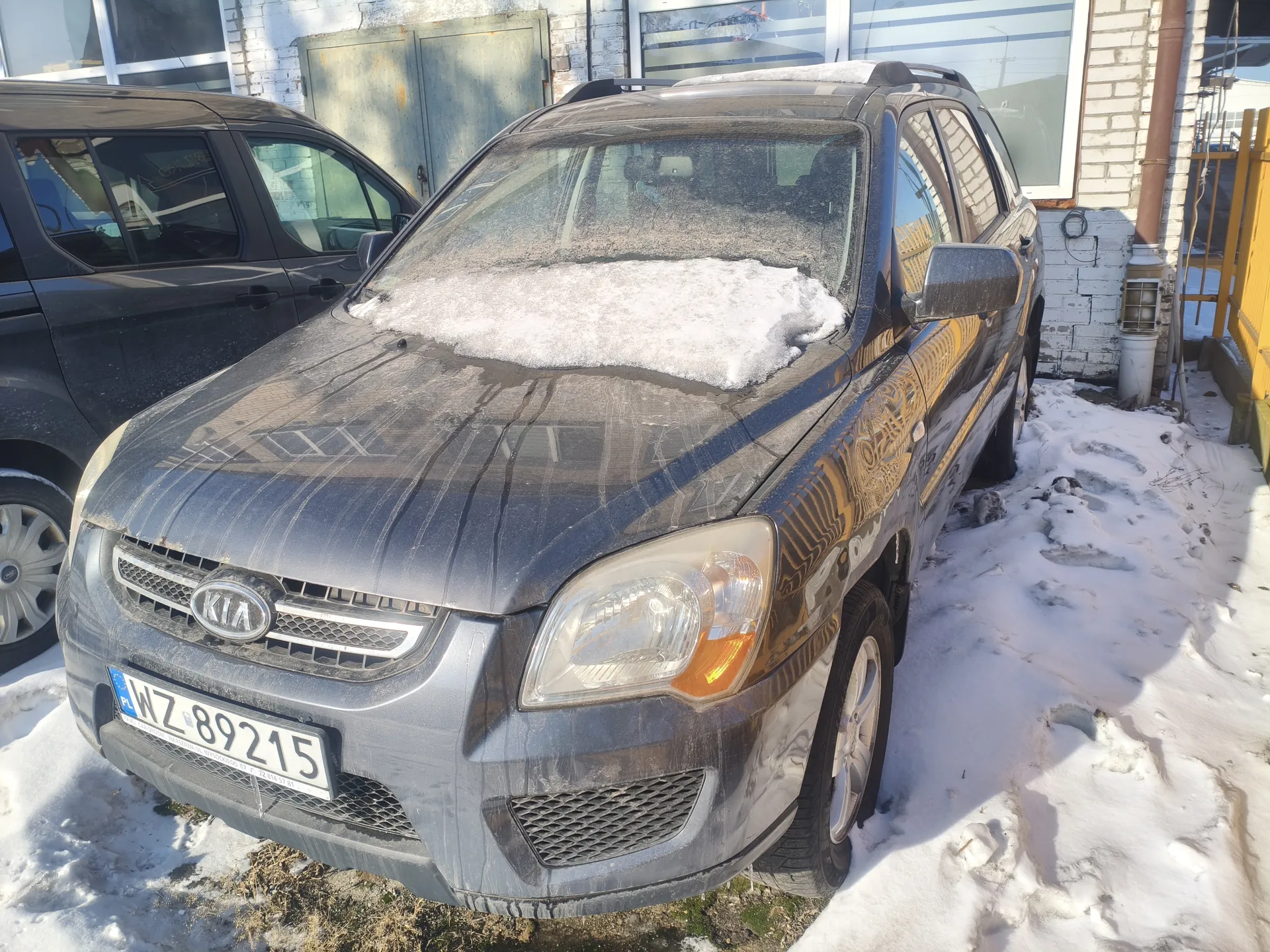 Ciemnoszary samochód marki Kia z tablicą rejestracyjną WZ 89215 zaparkowany na śniegu. Na masce widoczna warstwa śniegu i zabrudzenia. W tle budynek z białej cegły, metalowe drzwi oraz fragment drugiego ciemnego samochodu. Na tablicy rejestracyjnej widoczny niebieski pas z oznaczeniem PL.