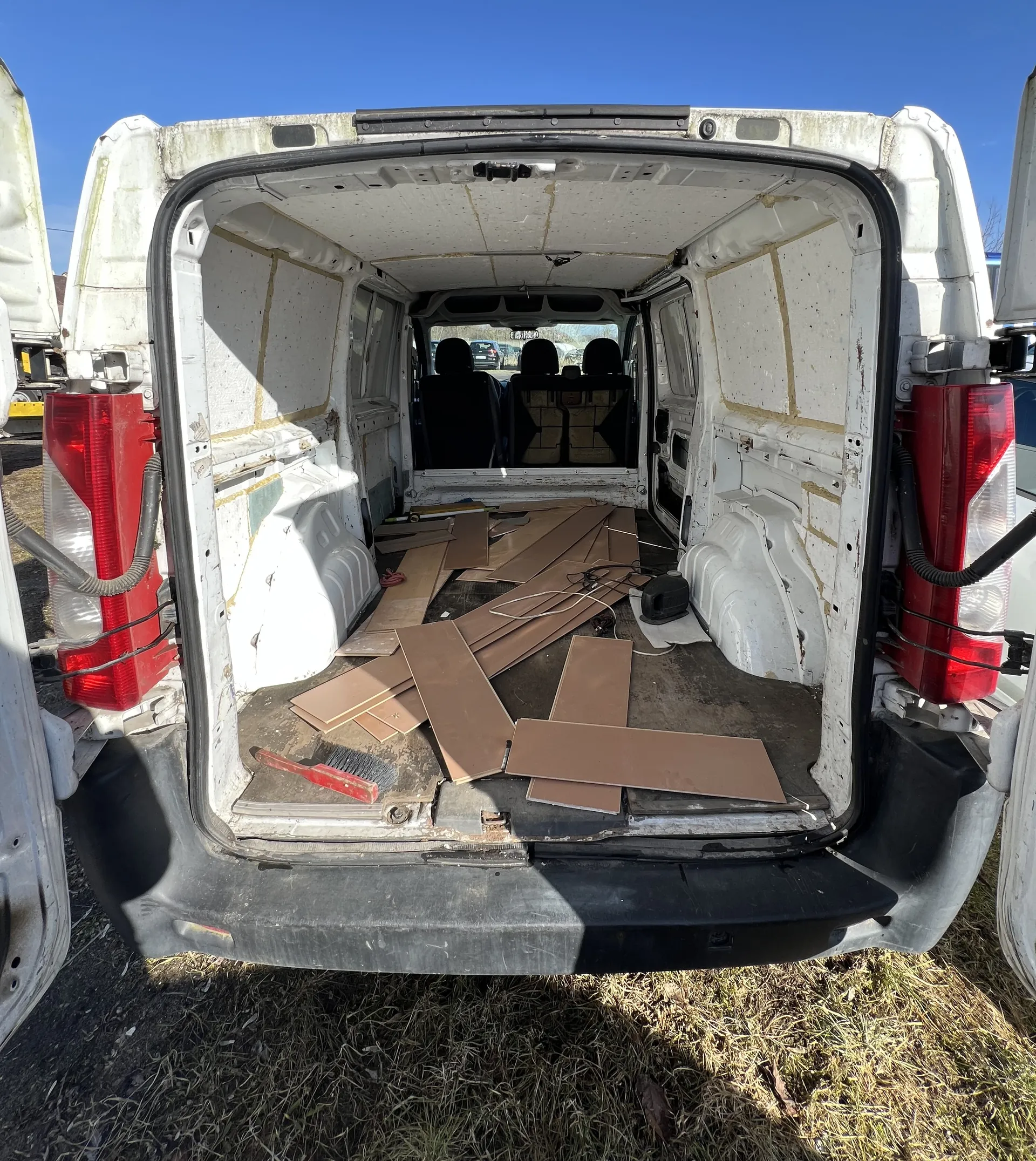 Widok z tyłu na otwarte drzwi białego furgonu. Wewnątrz furgonu leżą rozrzucone brązowe deski, czerwony pędzel oraz przewody. W tle widoczne są czarne siedzenia i niebieskie niebo. Furgon stoi na trawie, a jego zewnętrzne ściany mają ślady zużycia i zabrudzenia.