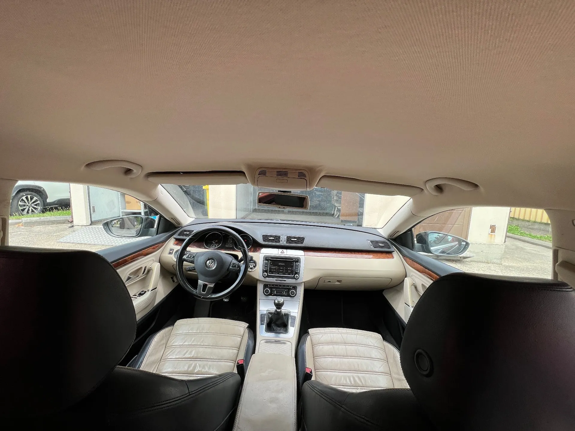 Wnętrze samochodu marki Volkswagen widziane z perspektywy tylnego siedzenia. Widoczna jest deska rozdzielcza w kolorze beżowym i brązowym, czarna kierownica z logo Volkswagena, konsola środkowa z lewarkiem zmiany biegów oraz dwa przednie fotele wykonane z beżowej i czarnej skóry. Przez przednią szybę widoczny jest fragment podwórka i zaparkowany biały samochód.