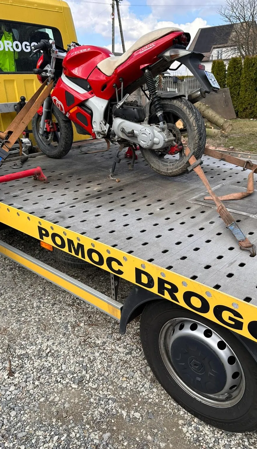 Czerwony motocykl marki FILERA z białymi i czarnymi elementami, zabezpieczony paskami na platformie holowniczej. Na żółto-czarnej krawędzi platformy widoczny napis 'POMOC DROGOWA'. W tle żółty pojazd z częściowo widocznym napisem 'DROG...' w oknie, domy, krzewy i niebo z chmurami. Podłoga platformy ma perforowane otwory, a pod nią żwirowa powierzchnia.