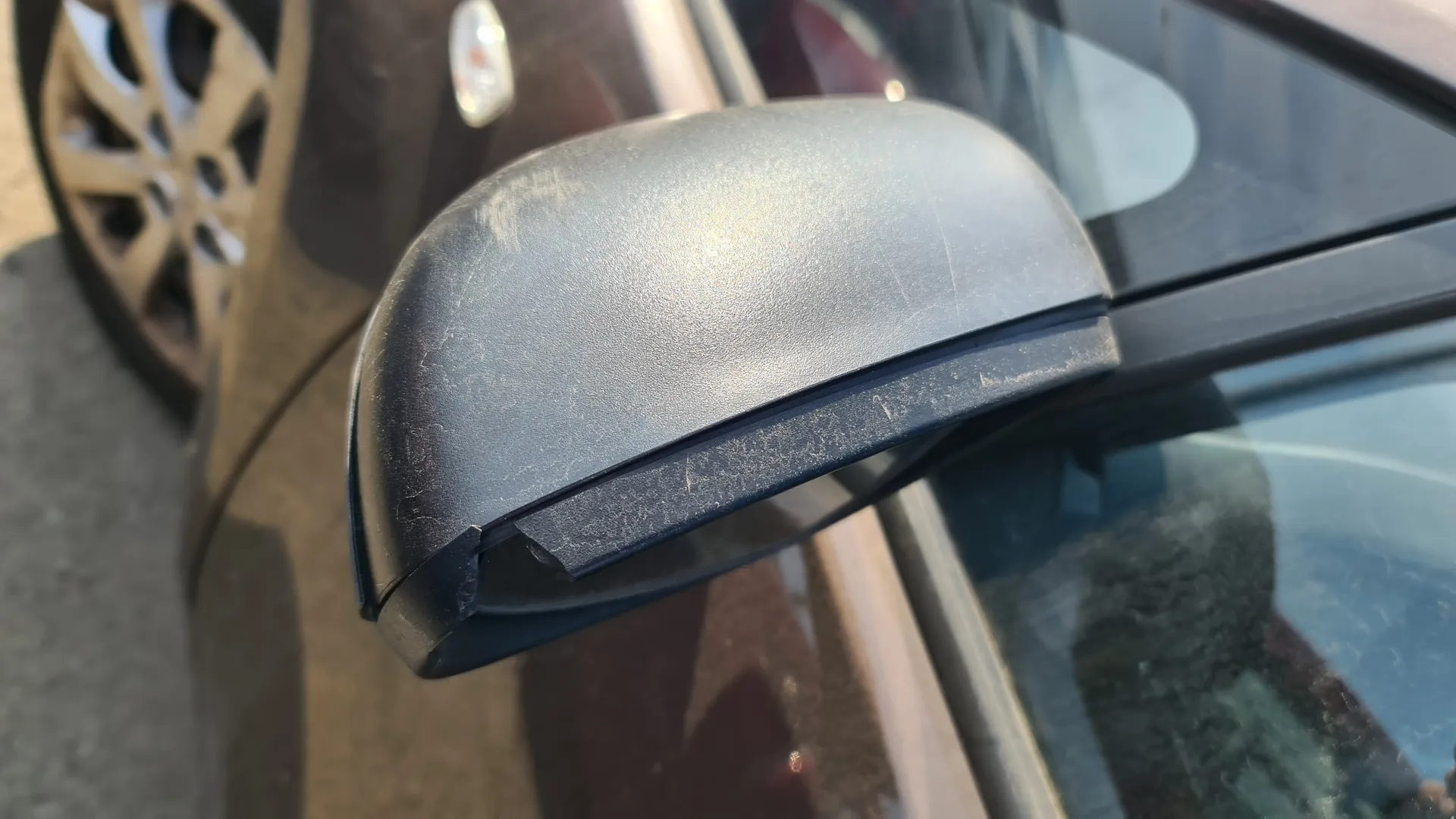 Zbliżenie na czarny boczny lusterko samochodu z widocznymi uszkodzeniami na dolnej krawędzi. W tle fragment ciemnego nadwozia samochodu, koło z srebrną felgą oraz asfaltowa powierzchnia. Lusterko ma lekkie zadrapania i uszkodzony plastik na krawędzi.