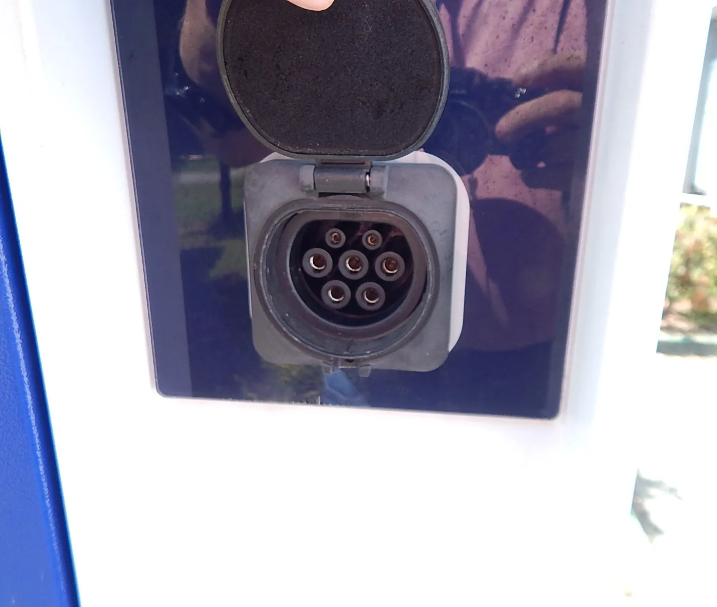 Zbliżenie na gniazdo ładowania pojazdu elektrycznego z otwartą, czarną klapką ochronną. Gniazdo jest okrągłe, posiada siedem pinów wewnątrz i jest osadzone w szarej obudowie na ciemnoniebieskim tle.