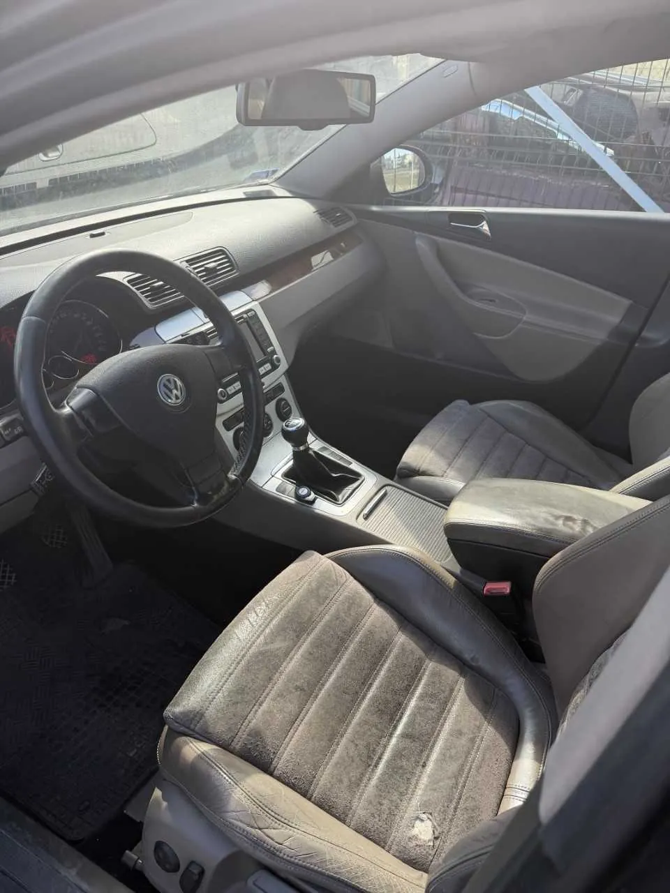 Wnętrze samochodu marki Volkswagen z widocznym kierownicą z logo VW, deską rozdzielczą, przyciskami sterującymi, dźwignią zmiany biegów oraz usłojonymi siedzeniami z czarnego materiału. W tle przez przednią szybę widoczny jest ogrodzenie i budynki. Wnętrze wydaje się nieco zabrudzone, a siedzenie kierowcy ma widoczne uszkodzenie na powierzchni.