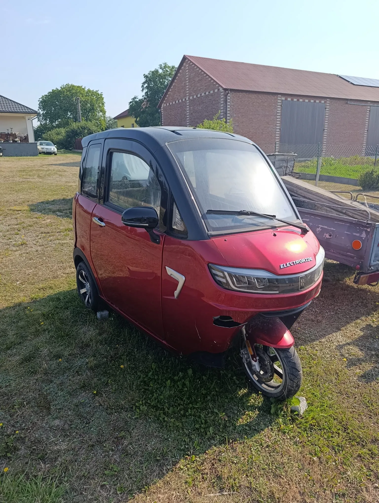 Mały, czerwony samochód elektryczny marki Electroride zaparkowany na trawiastym terenie. Pojazd ma czarny dach i czarne ramki szyb. W tle widoczny jest ceglany budynek gospodarczy, zielone drzewa oraz fragment innego białego samochodu w oddali.