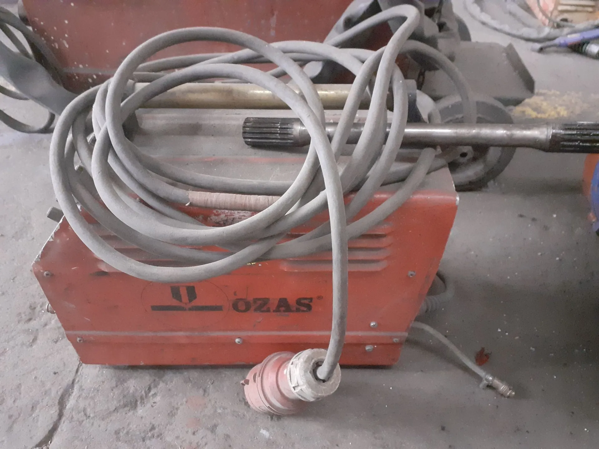 Pomarańczowa maszyna warsztatowa z napisem 'OZAS', na której leży zwinięty, szary kabel elektryczny z wtyczką. W tle widoczne są elementy mechaniczne, w tym metalowy wał z ząbkami.
