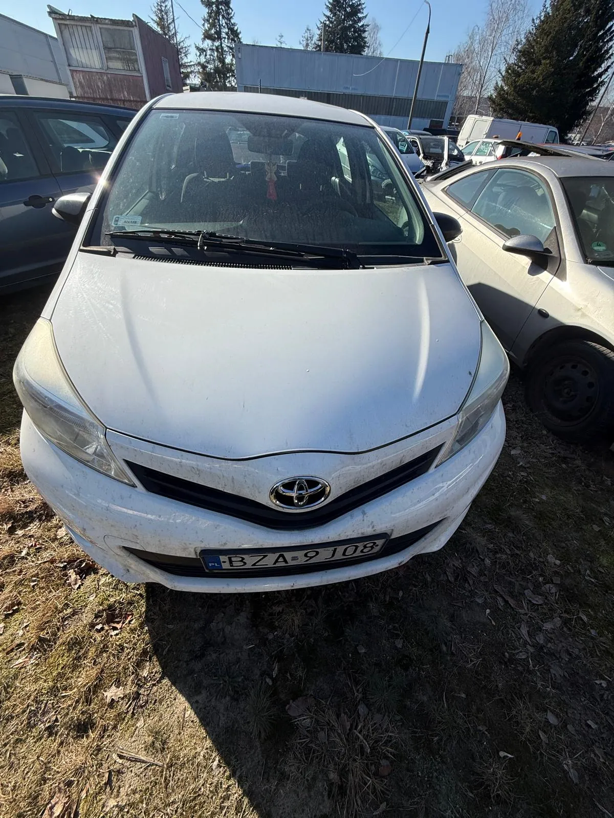 Biały samochód marki Toyota z tablicą rejestracyjną PL BZA 9J08, zaparkowany na trawie z suchymi liśćmi. W tle widoczne są inne pojazdy, budynki przemysłowe, drzewa i jasne niebo. Samochód ma zabrudzony nadwoziem, a na przedniej szybie widoczne są wycieraczki i czerwony przedmiot wiszący wewnątrz.
