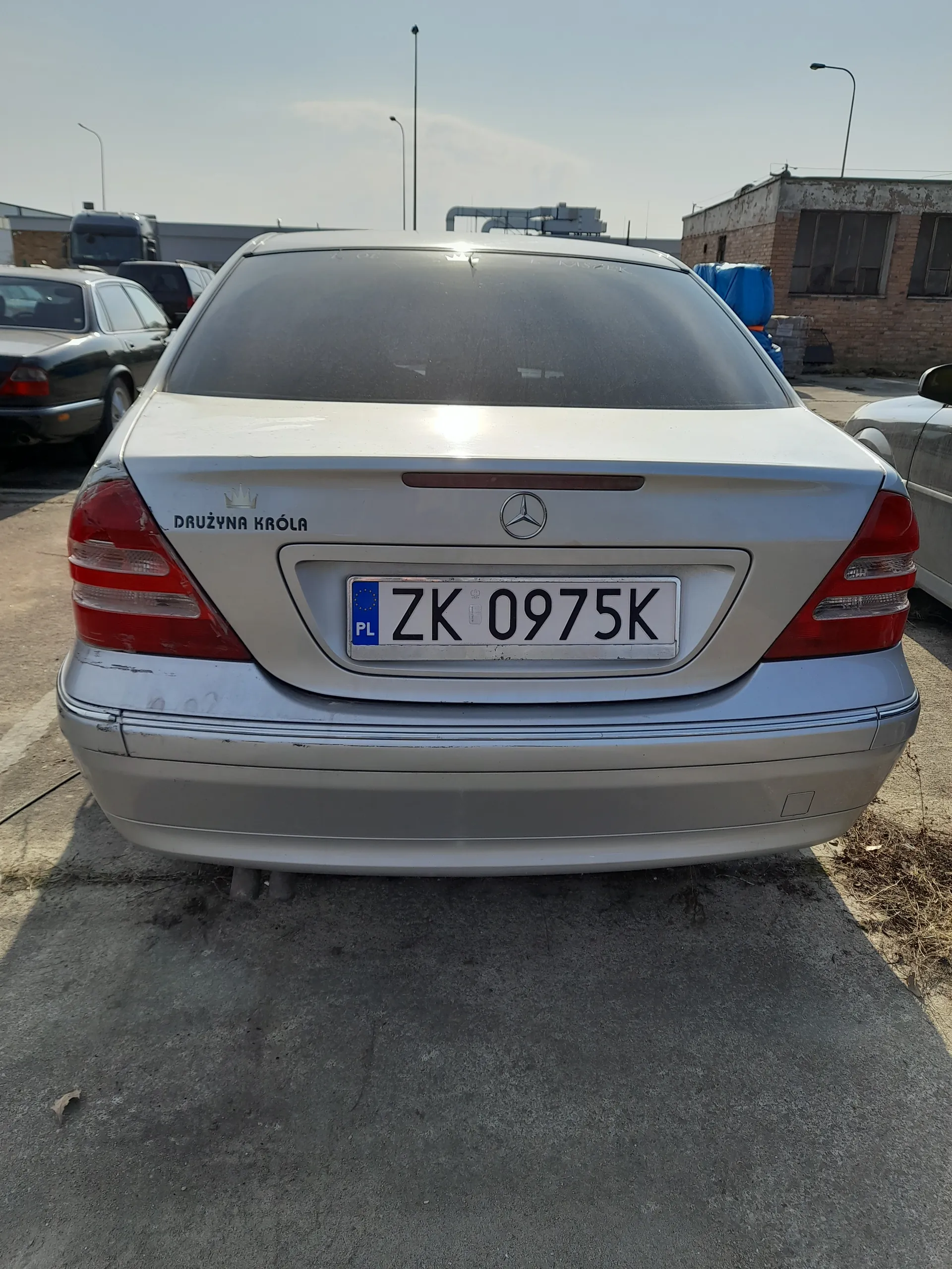 Tył srebrnego samochodu marki Mercedes-Benz z tablicą rejestracyjną ZK 0975K z niebieskim pasem PL po lewej stronie. Na bagażniku widoczny napis DRUŻYNA KRÓLA oraz logo Mercedes-Benz. W tle budynki, inne zaparkowane pojazdy, latarnie uliczne i asfaltowa powierzchnia. Niebo jest widoczne po lewej stronie obrazu.
