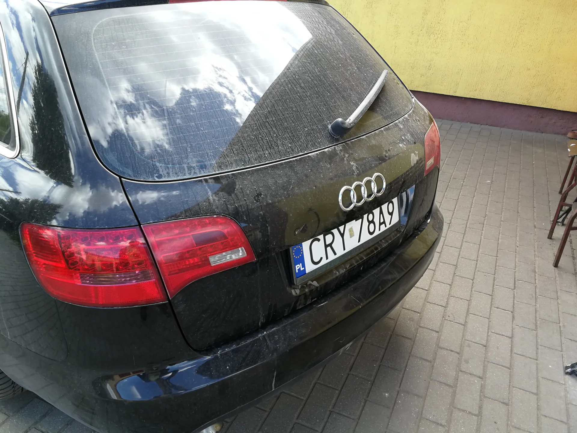 Tył czarnego samochodu marki Audi z widocznym logo, zabrudzonym nadwoziem i tablicą rejestracyjną PL CRY 78A9. W tle żółta ściana z brązowym pasem, brukowana powierzchnia oraz fragmenty czerwonych krzeseł po prawej stronie. Na szybie tylnej odbijają się chmury i drzewa.