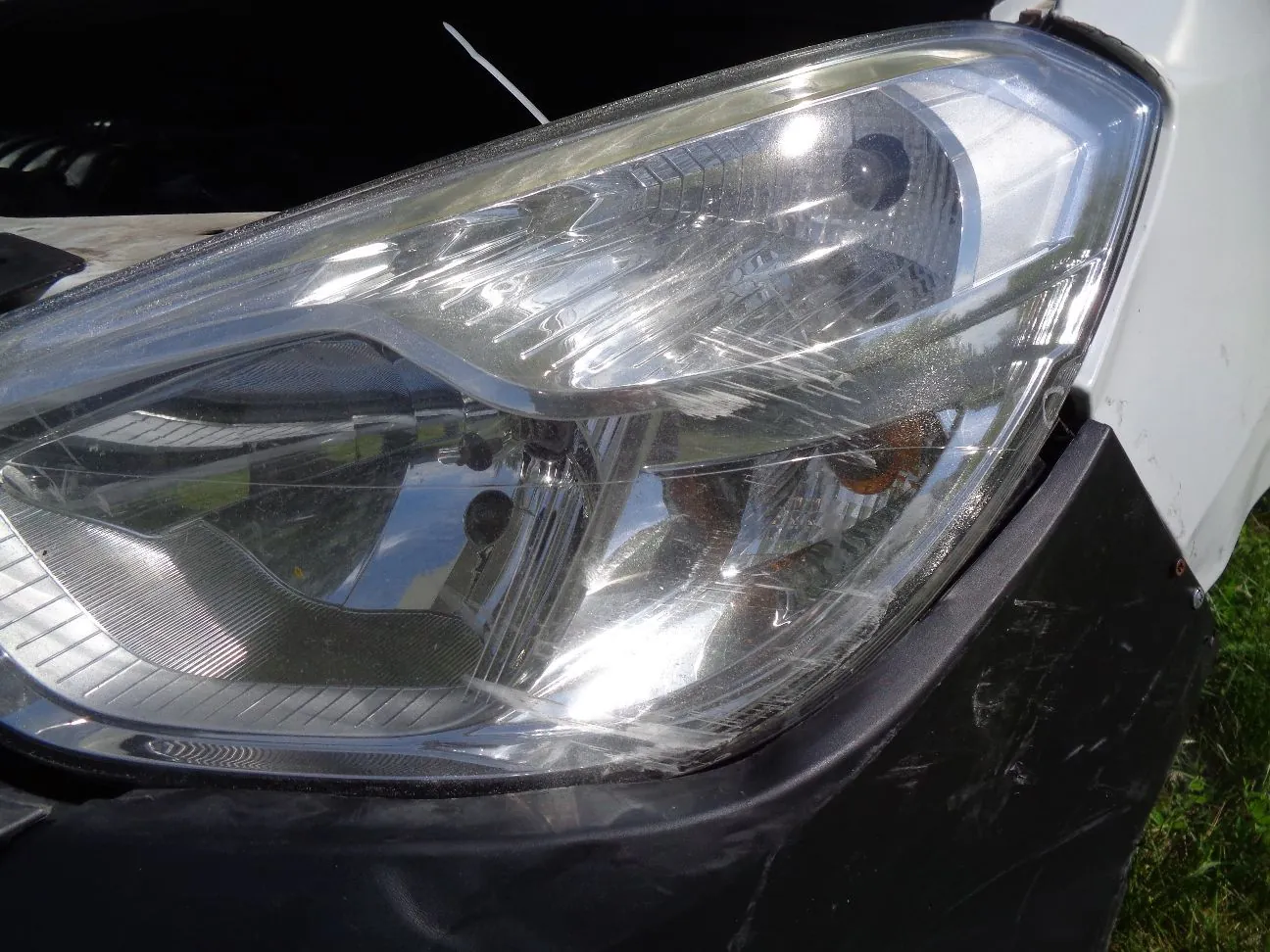 Zbliżenie na przedni reflektor samochodu w kolorze białym. Przezroczysta obudowa lampy jest porysowana i zmatowiona. Poniżej reflektora widoczny jest czarny, plastikowy element zderzaka z widocznymi zarysowaniami.