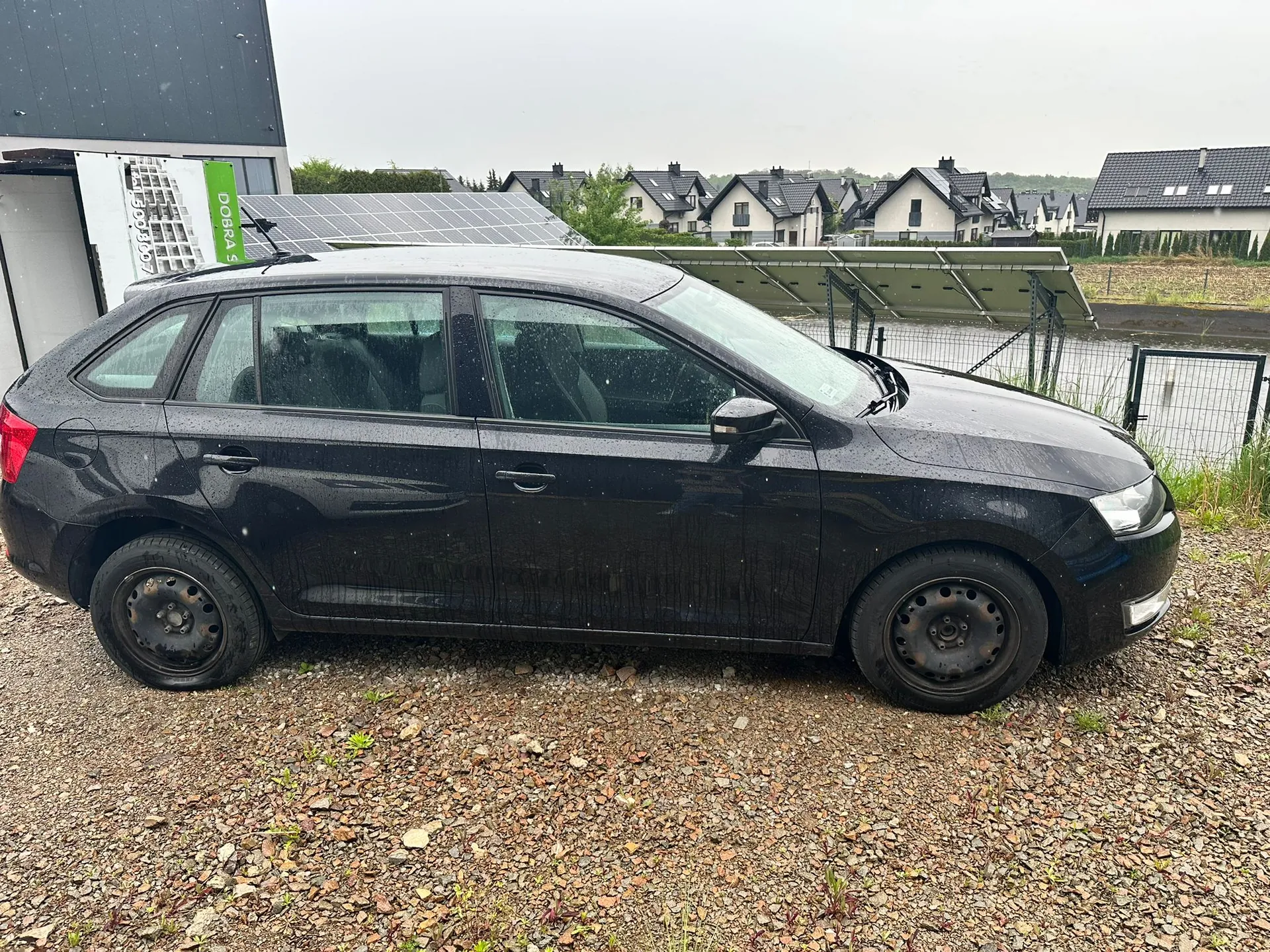 Czarny samochód typu hatchback zaparkowany na żwirowym podłożu. Na karoserii i szybach widoczne krople deszczu. W tle domy z ciemnymi dachami, panele słoneczne oraz zielony znak z napisem DOBRA 5. Po lewej stronie budynek z ciemnym elewacją, a na niebie pochmurna pogoda. W oddali siatka ogrodzeniowa i zielone krzewy.