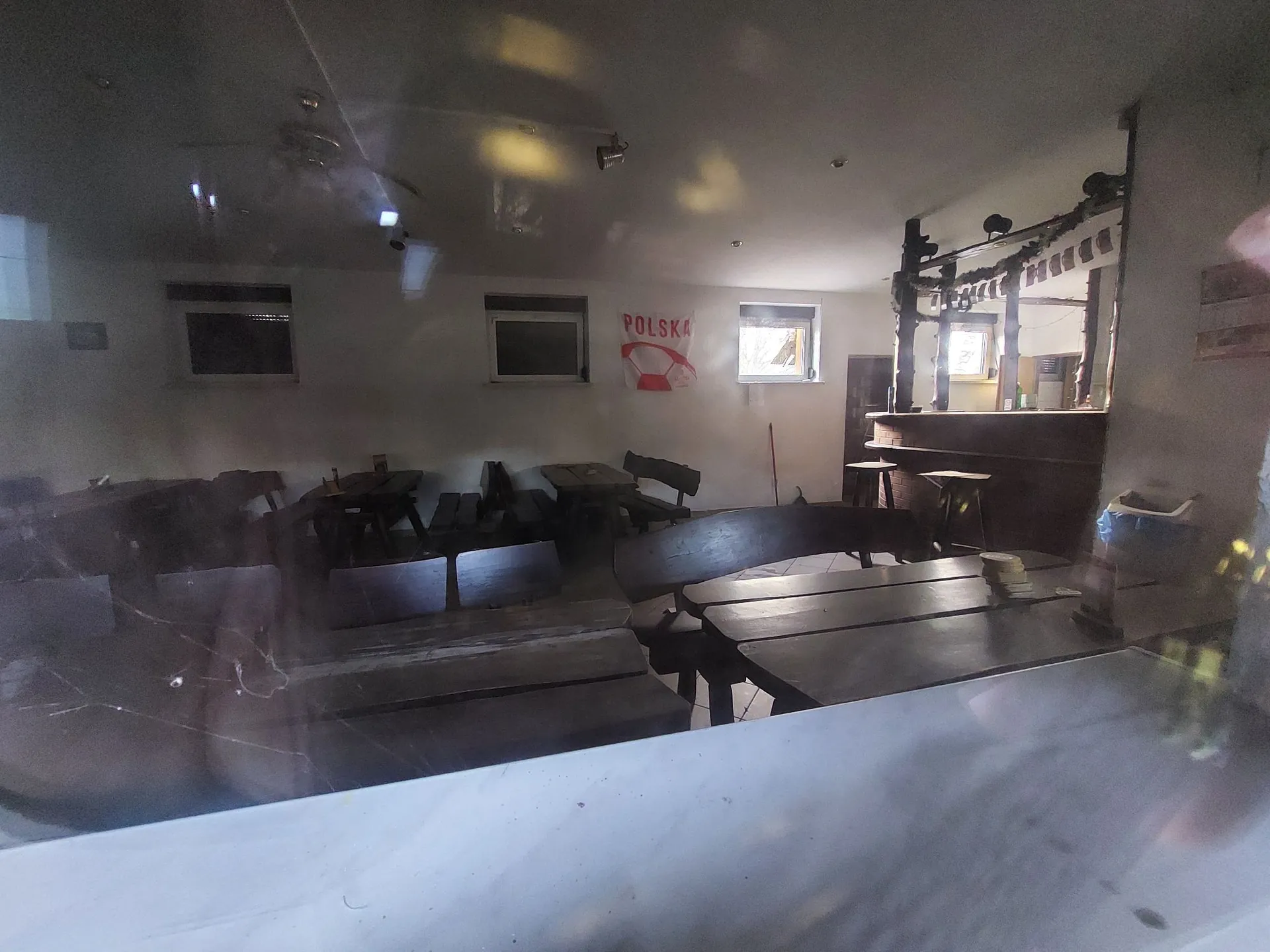 Wnętrze ciemnego, opuszczonego pomieszczenia przypominającego bar lub restaurację. W pomieszczeniu znajdują się drewniane stoły i ławy. Na białej ścianie w tle wisi flaga Polski z napisem „POLSKA”. Po prawej stronie widoczny jest ciemny bar z hokerami. Zdjęcie zostało wykonane przez szybę, na której widoczne są odbicia i zabrudzenia.