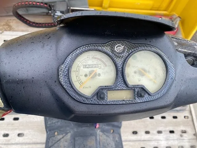 Zbliżenie na czarny deski rozdzielczej pojazdu z dwoma okrągłymi wskaźnikami. Lewy wskaźnik to prędkościomierz z zakresem do 80 km/h, na wyświetlaczu cyfrowym widoczny przebieg 14996. Prawy wskaźnik ma nieczytelne oznaczenia. Między wskaźnikami znajduje się mały wyświetlacz cyfrowy. Na desce rozdzielczej widoczny jest logo oraz wzór podobny do włókna węglowego. W tle żółta konstrukcja i metalowa kratka. Na powierzchni deski rozdzielczej krople wody.