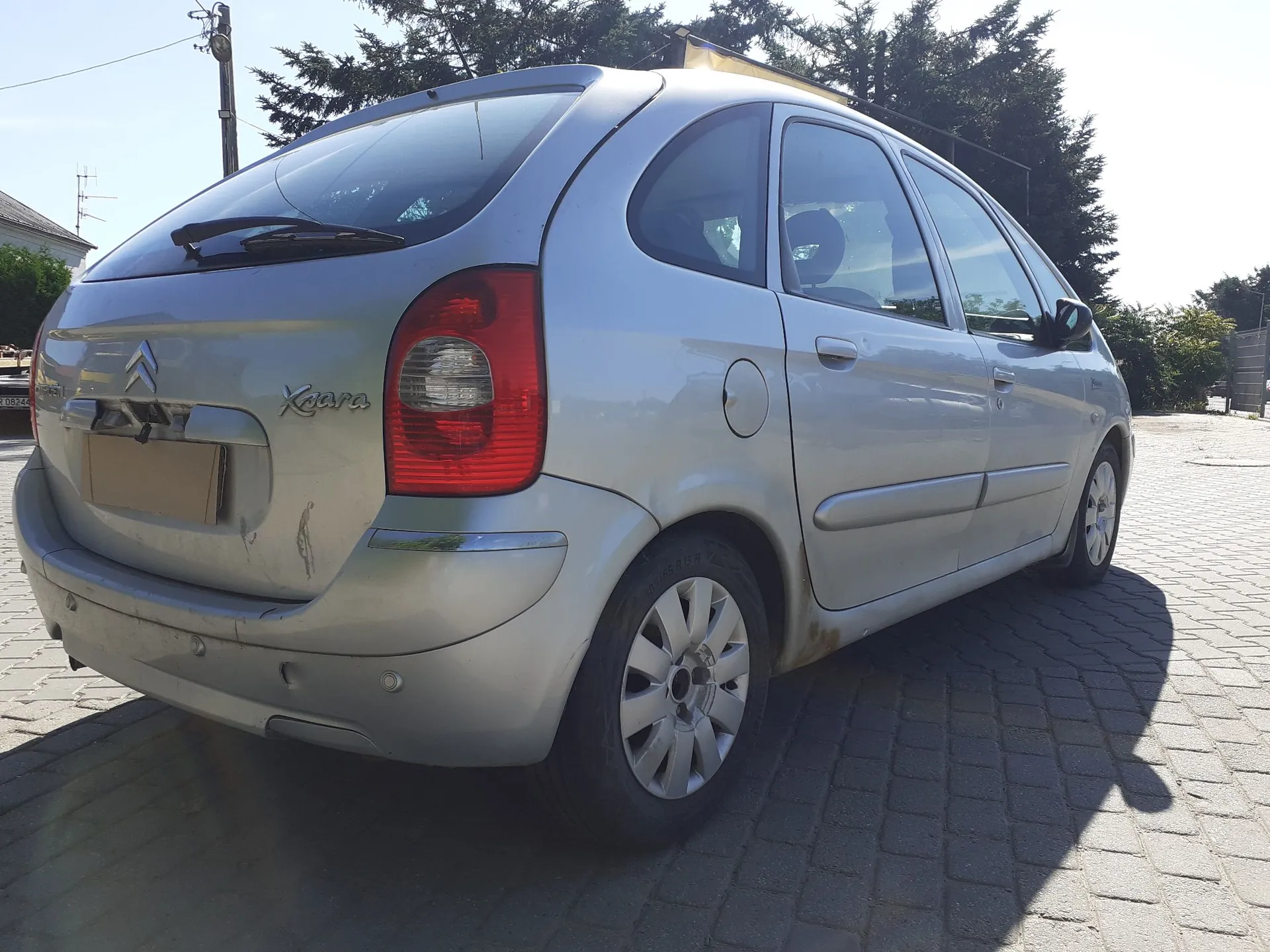 Srebrny samochód marki Citroën Xsara w nadwoziu hatchback, zaparkowany na kostce brukowej. Widok z tyłu i z boku pojazdu. Na klapie bagażnika widoczne jest logo Citroëna oraz napis 'Xsara'. Samochód ma srebrne felgi aluminiowe.