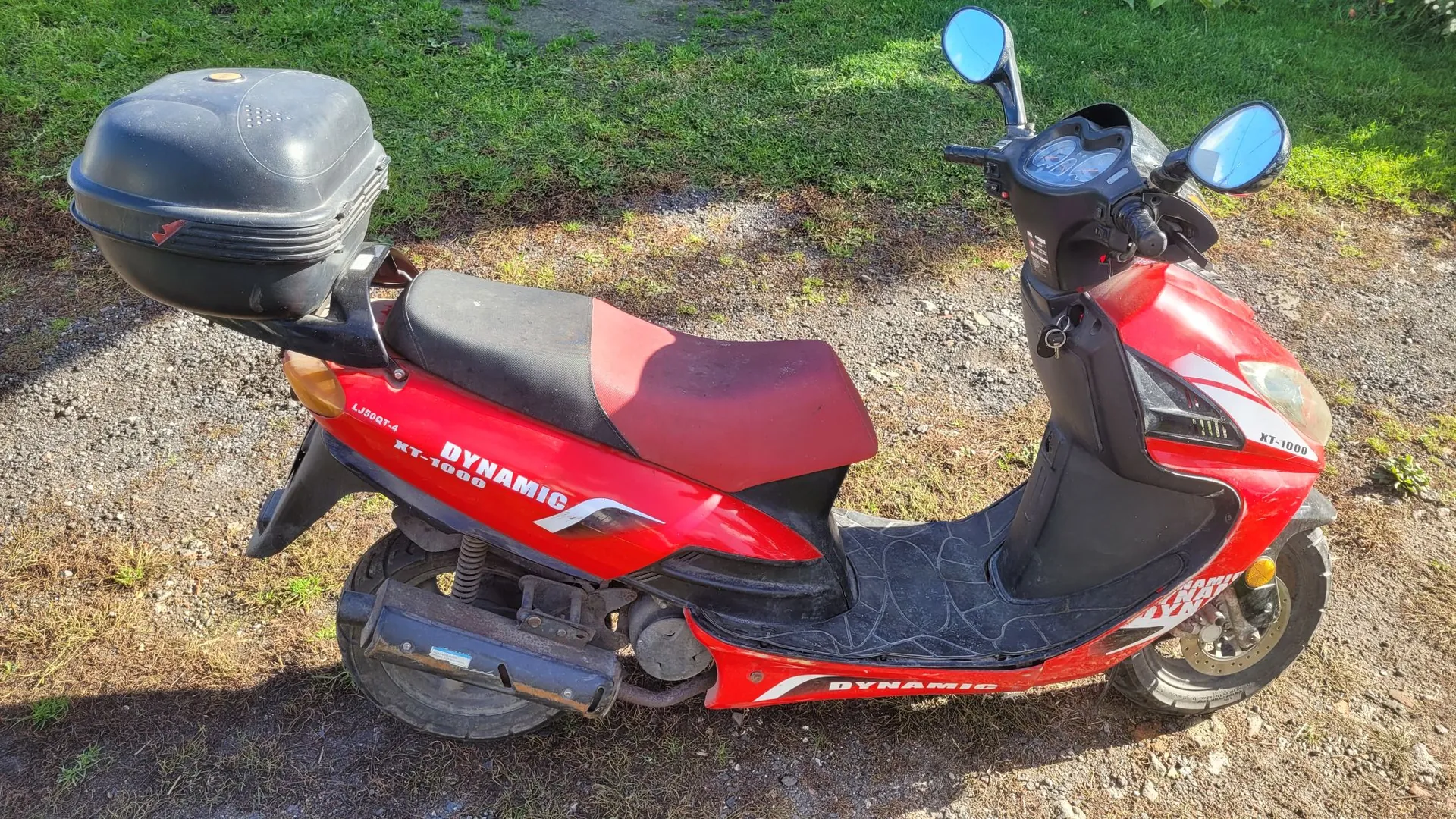 Czerwony skuter marki Dynamic, model XT-1000, zaparkowany na piaszczysto-trawiastej nawierzchni. Pojazd posiada czarny kufer zamykany na klucz zamontowany z tyłu, czerwone siedzenie oraz czarne elementy plastikowe. Na boku skutera widnieją białe napisy 'DYNAMIC' oraz 'XT-1000'.