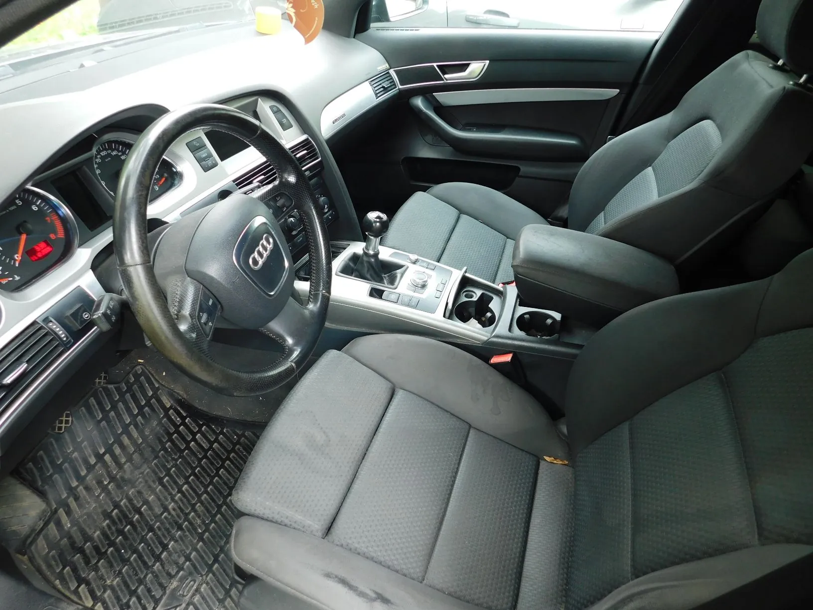 Wnętrze samochodu marki Audi widziane z perspektywy pasażera. Widoczna jest czarna kierownica z logo Audi, deska rozdzielcza, automatyczna skrzynia biegów, dwa przednie fotele z ciemnoszarej tkaniny oraz gumowa mata na podłodze po stronie kierowcy.
