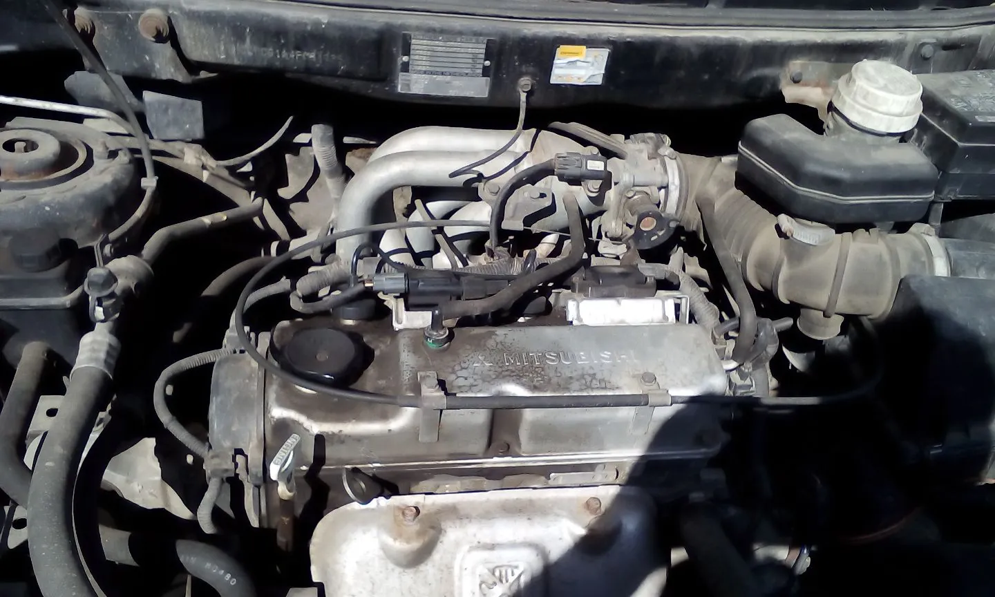 Zbliżenie na komorę silnika samochodu. Widoczny jest metalowy blok silnika z napisem MITSUBISHI na pokrywie zaworów, układ dolotowy, przewody elektryczne oraz zbiornik płynu z białką zakrętką po prawej stronie.