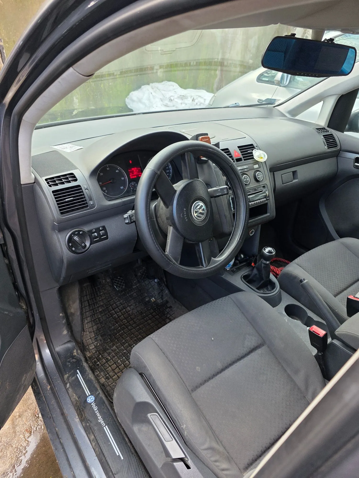 Wnętrze samochodu marki Volkswagen widziane z perspektywy otwartych drzwi kierowcy. Widoczna jest czarna kierownica z logo marki, deska rozdzielcza z analogowymi wskaźnikami prędkości i obrotów, otwory nawiewów oraz konsola środkowa z lewarkiem zmiany biegów. W kadrze znajdują się dwa przednie fotele z ciemnoszarej tkaniny oraz gumowa mata na podłodze. Na progu samochodu widnieje napis 'volkswagen'.
