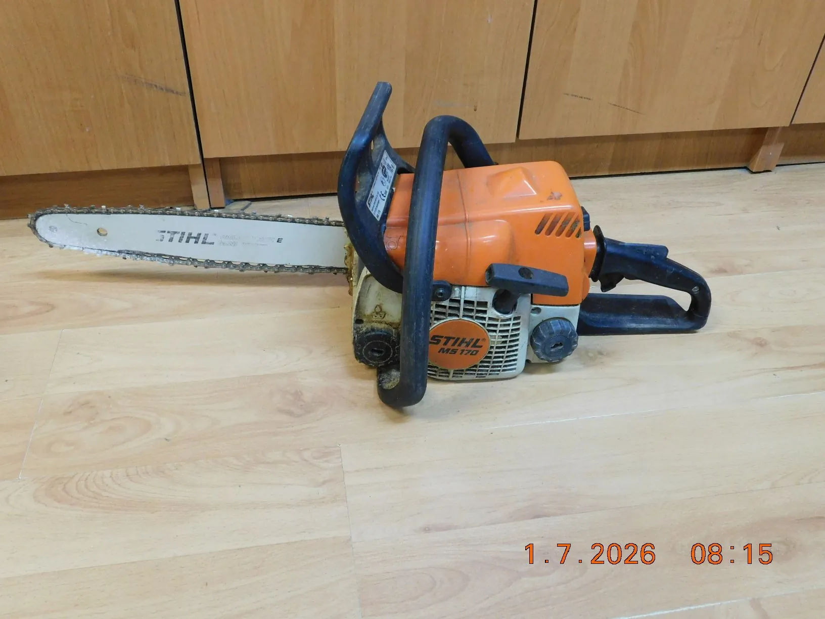 Pomarańczowo-biała piła łańcuchowa marki STIHL model MS 170, leżąca na jasnej drewnianej podłodze przed drewnianymi szafkami. Na prowadnicy piły widoczny jest napis STIHL, a na obudowie silnika napis STIHL MS 170. W prawym dolnym rogu zdjęcia znajduje się pomarańczowy znacznik czasu: 1.7.2026 08:15.