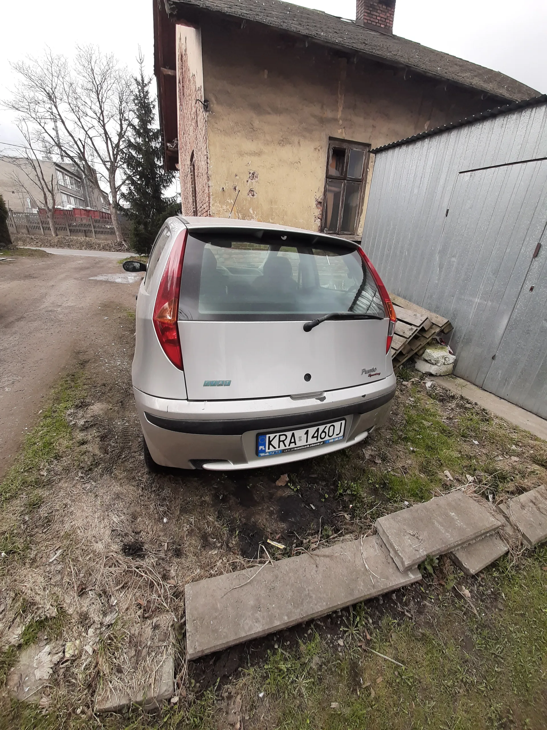 Srebrny samochód zaparkowany na podwórku obok budynku o żółtej ścianie i metalowym dachu. Widoczna tablica rejestracyjna KRA 1460J z niebieskim pasem z napisem PL. Wokół trawa, betonowe płyty i fragmenty muru. W tle drzewa i inne budynki. Samochód ma czerwone światła tylna i czarny wycieraczka na tylnej szybie.