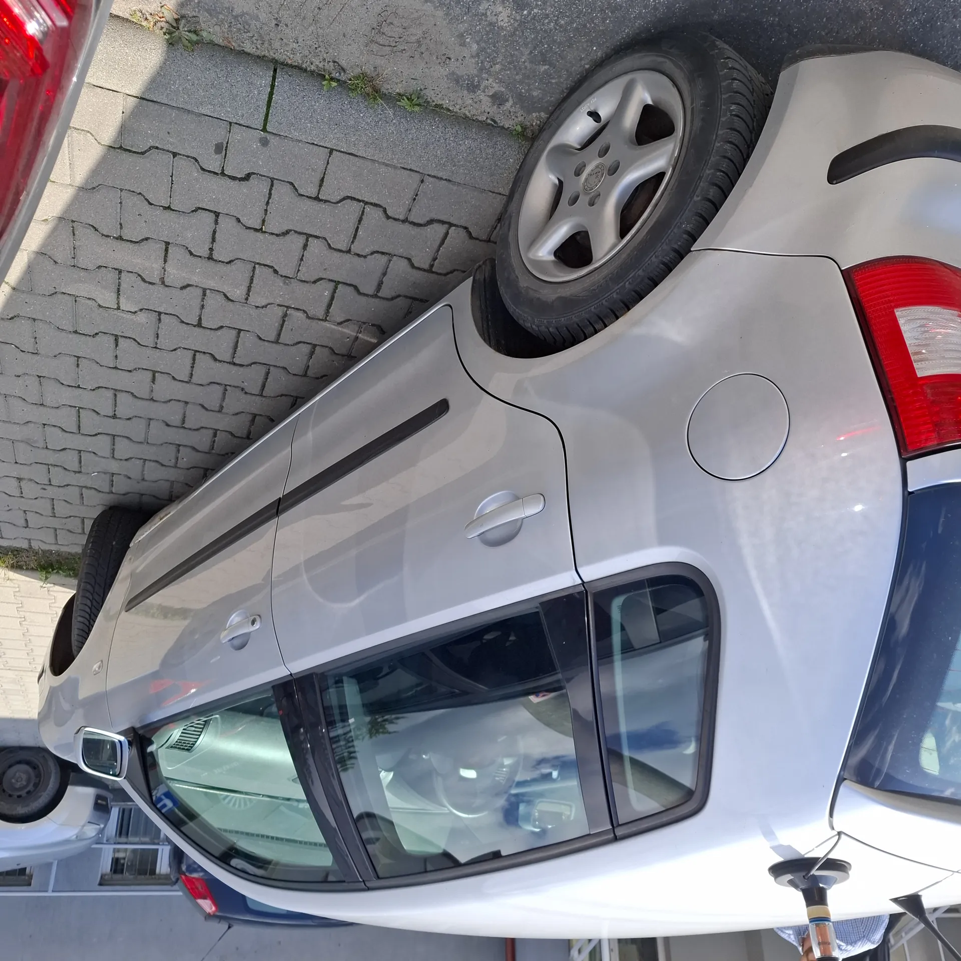 Srebrny samochód osobowy zaparkowany na brukowanej powierzchni. Widoczne są boczne drzwi, szyby, lusterko boczne oraz tylna część z czerwonym światłem. W tle inne pojazdy, budynek z oknami i brukowane chodniki. Na chodniku rosną pojedyncze rośliny.