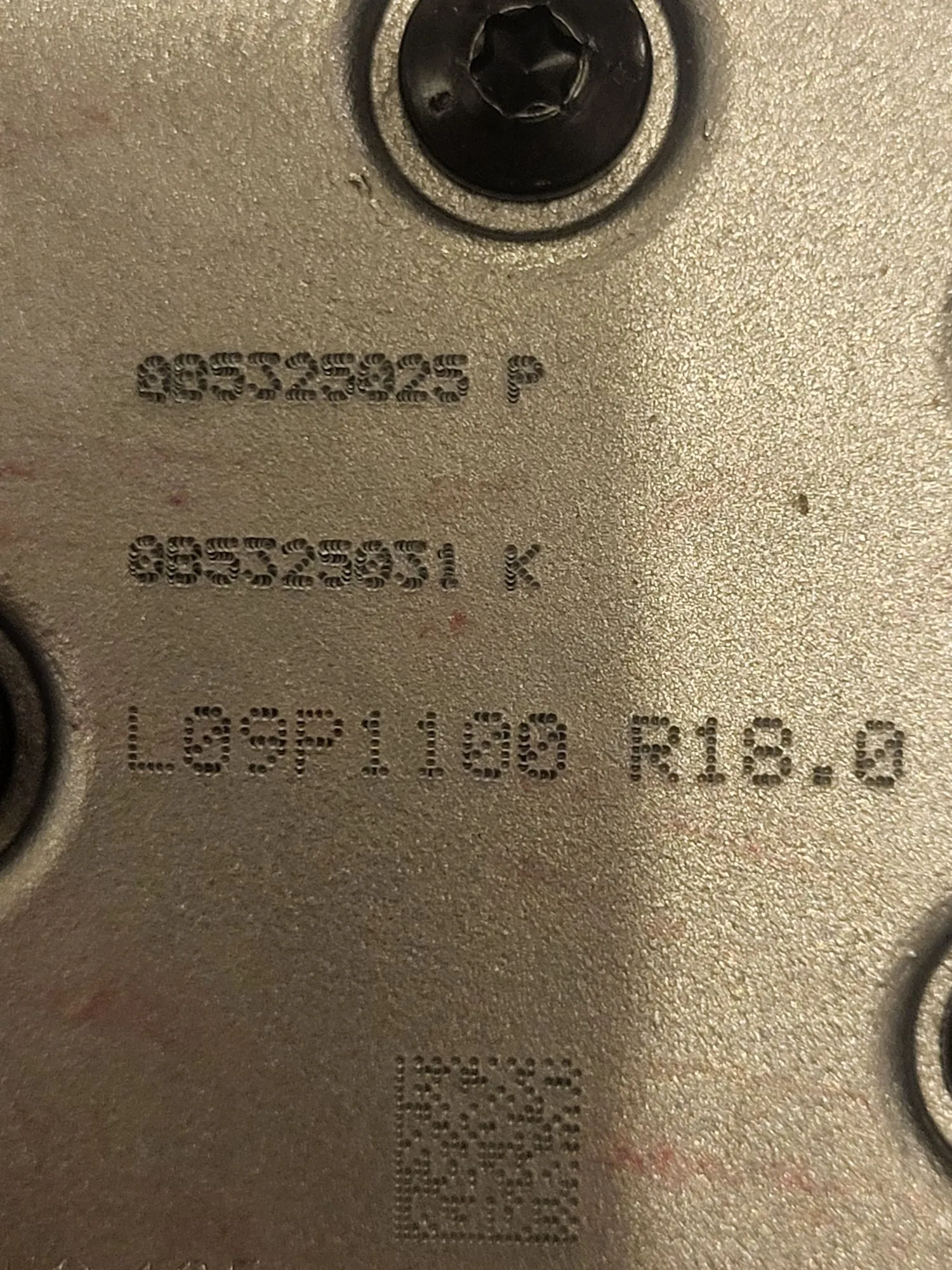 Zbliżenie na metalową powierzchnię z grawerowanymi numerami seryjnymi i kodem QR. Widoczne są trzy linie tekstu: '885325925 P', '885325931 K' oraz 'L09P1100 R18.0'. W górnej części znajduje się czarna śruba z gniazdem imbusowym, a na dole kwadratowy kod QR.