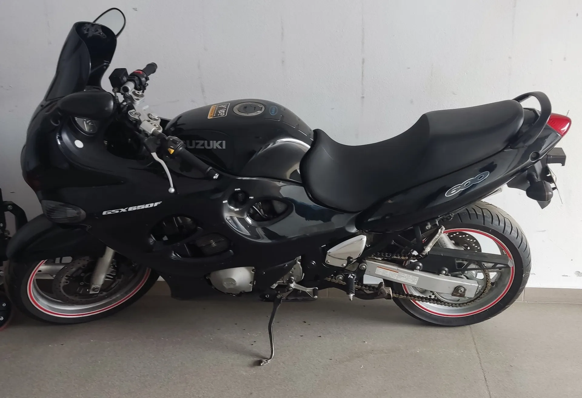 Czarny motocykl marki Suzuki model GSX650F z widocznym napisem 600 na boku. Motocykl posiada czarne siedzenie, czerwone pasy na obręczach kół, przednią szybę oraz czerwone światło tylnego. Na zbiorniku widoczny jest napis SUZUKI, a na boku napis GSX650F. Motocykl stoi na betonowej podłodze przy szarej ścianie. Widoczne są również elementy mechanizmu, łańcuch i podstawa.