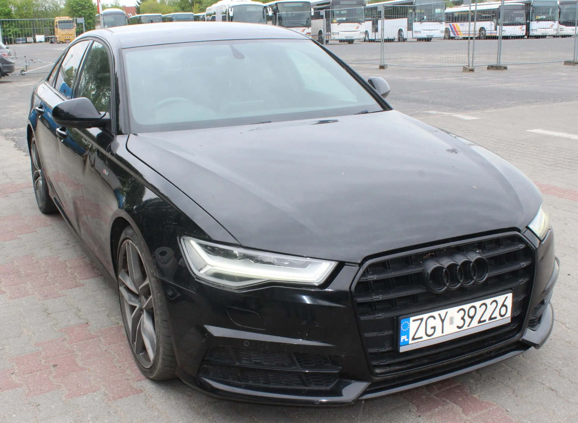 Czarny samochód marki Audi zaparkowany na kostce brukowej. Widoczny jest przód i lewy bok pojazdu. Samochód ma tablicę rejestracyjną o numerze ZGY 39226. W tle widać parking z zaparkowanymi białymi autobusami oraz żółty pojazd w oddali.