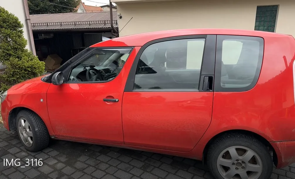 Czerwony samochód osobowy typu hatchback zaparkowany na brukowanej powierzchni. Widok z boku, widać drzwi, okna i koła. W tle budynek z beżową ścianą, zielonym oknem i garażem z przedmiotami wewnątrz. Po lewej stronie krzew. W lewym dolnym rogu widoczny napis "IMG_3116".