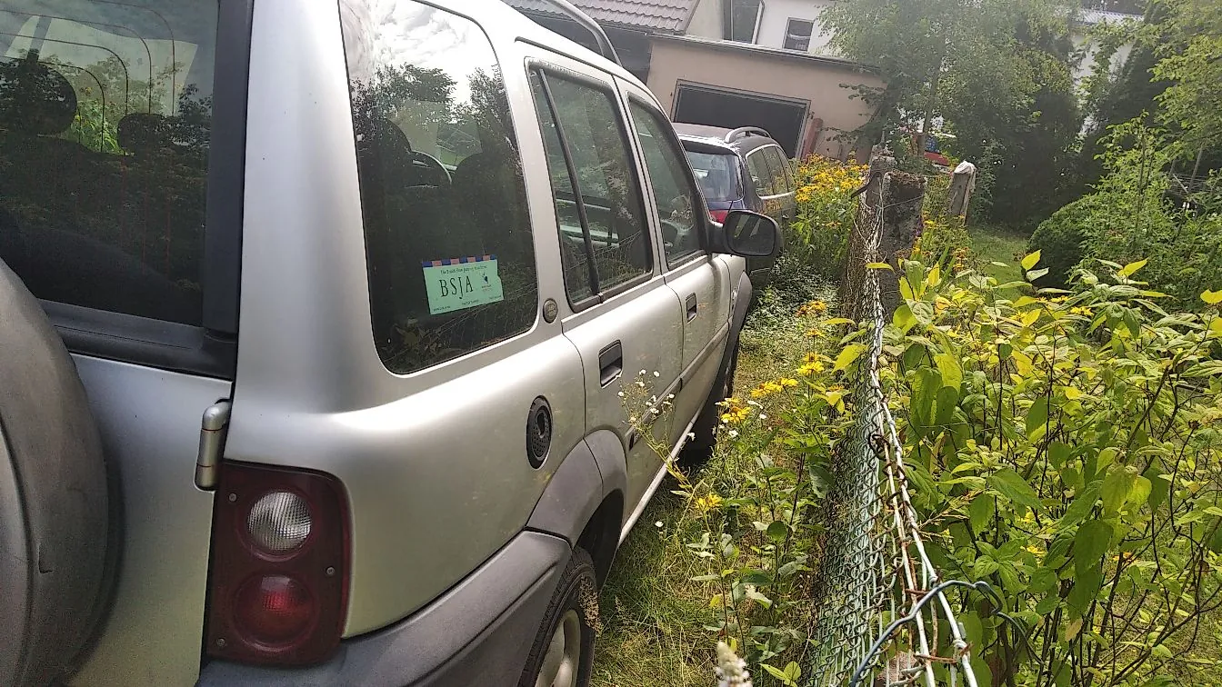 Srebrny samochód typu SUV zaparkowany obok zielonego ogrodzenia z siatki i gęstych roślin. Na tylnej szybie samochodu naklejona jest niebieska naklejka z białym tekstem 'BSJA'. W tle widoczny jest inny samochód oraz fragment budynku z garażem.