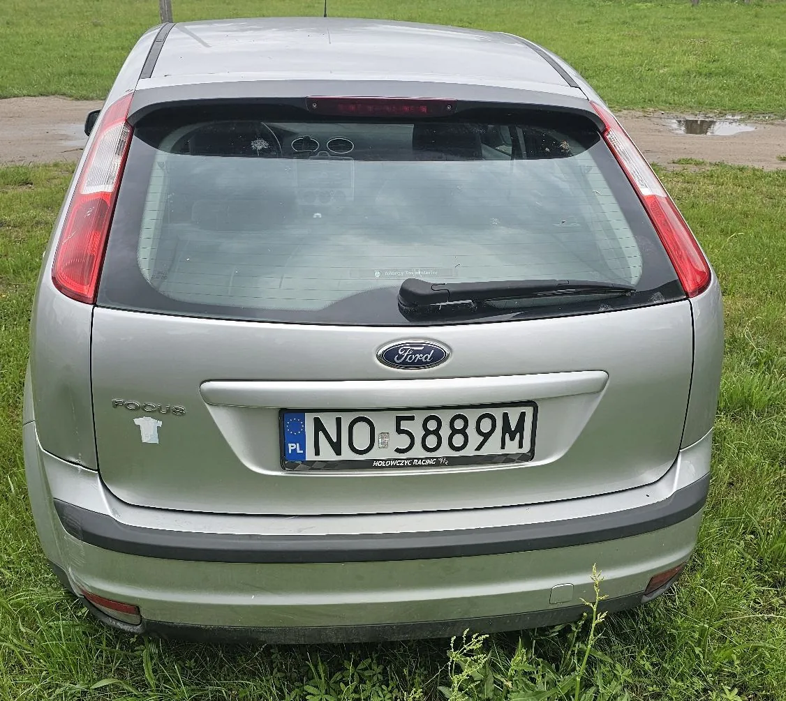 Tylny widok srebrnego samochodu marki Ford Focus zaparkowanego na trawie. Na klapie bagażnika znajduje się logo Ford oraz napis FOCUS. Samochód posiada polską tablicę rejestracyjną o numerze NO 5889M, a pod nią widnieje napis HOLOWCZYC RACING 1/2.