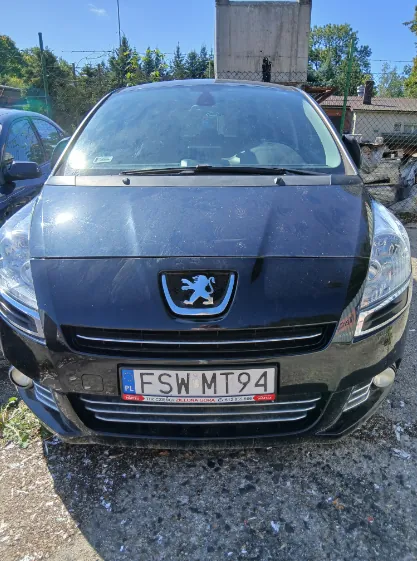 Widok z przodu na czarny samochód marki Peugeot. Na masce widoczne jest logo lwa w srebrnej ramce. Samochód posiada polską tablicę rejestracyjną o numerze FSW MT94. Pojazd stoi na żwirowym podłożu na tle zielonych drzew i betonowej konstrukcji.