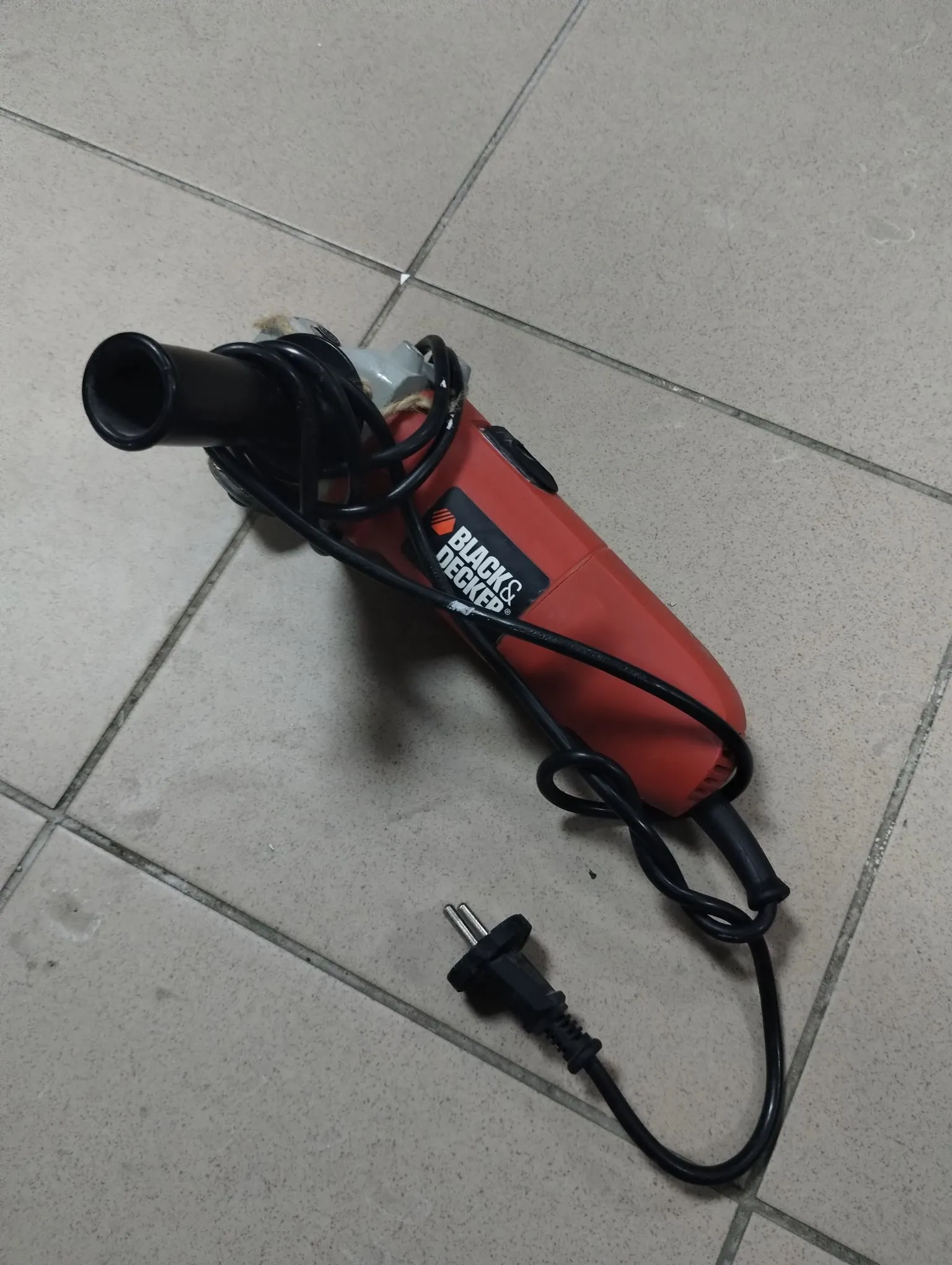 Pomarańczowa pistoletowa opalarka marki Black & Decker z czarnym kablem zasilającym i wtyczką, leżąca na jasnoszarej płytkowej podłodze.