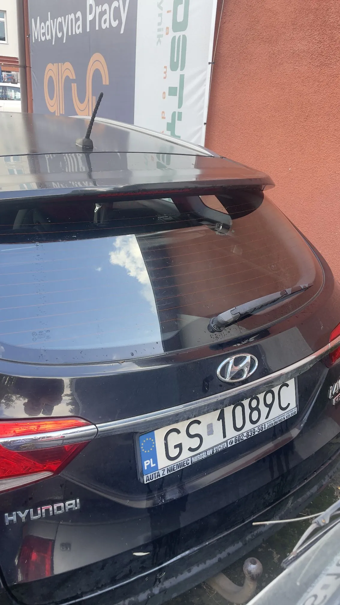 Tylna część czarnego samochodu marki Hyundai. Widoczna jest tylna szyba z wycieraczką, spojler oraz tablica rejestracyjna o numerze GS 1089C. Poniżej numeru rejestracyjnego znajduje się napis: 'PL AUTA Z NIEMIEC MIROSŁAW SYCZYŁA 602-839-561'. W tle widoczny jest fragment czerwonej ściany budynku oraz baner z napisem 'Medycyna Pracy'.