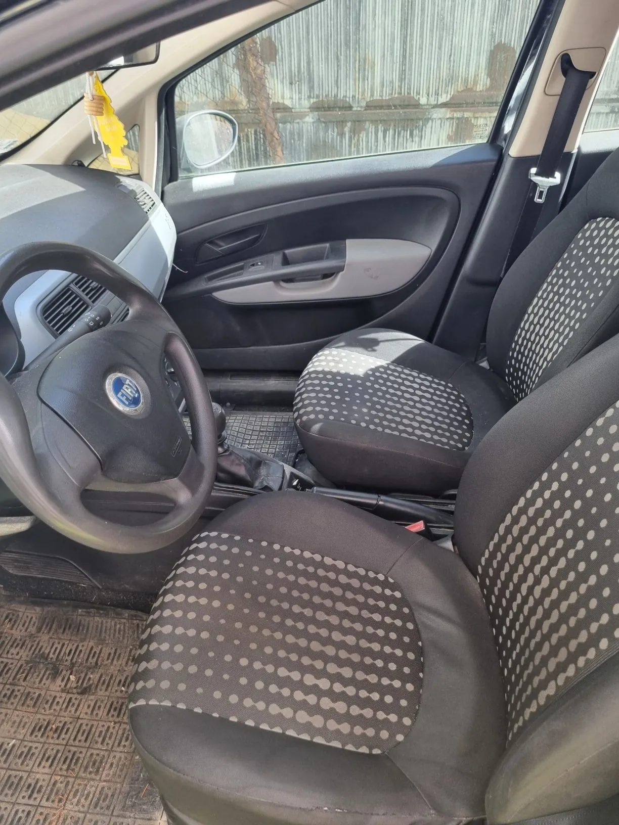 Wnętrze samochodu z perspektywy kierowcy. Widoczna jest czarna kierownica z logo marki Fiat, dwa przednie fotele z czarną tapicerką w drobne, jasne kropki, panel drzwi kierowcy oraz fragment deski rozdzielczej. Przez szybę widoczny jest fragment ogrodzenia z metalowych płyt.