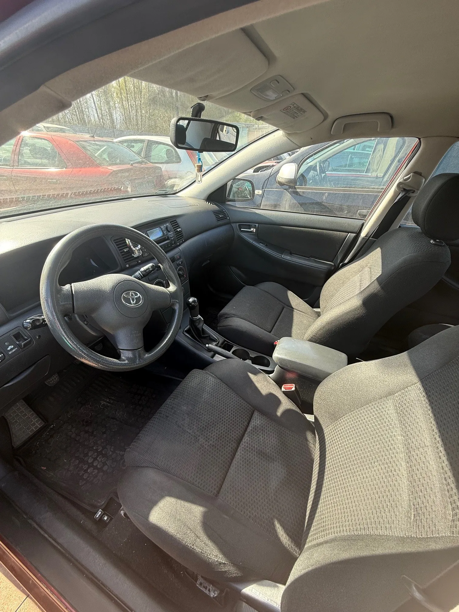 Wnętrze samochodu marki Toyota z widocznym kierownicą z logo, szarymi siedzeniami tapicerowanymi tkaniną, czarną deską rozdzielczą i centralną konsolą z dźwignią zmiany biegów. Przez przednią szybę widać zaparkowane samochody, w tym czerwony i biały. W lusterku wstecznym widoczny jest fragment otoczenia. Słońce oświetla wnętrze, rzucając cienie na siedzenia.