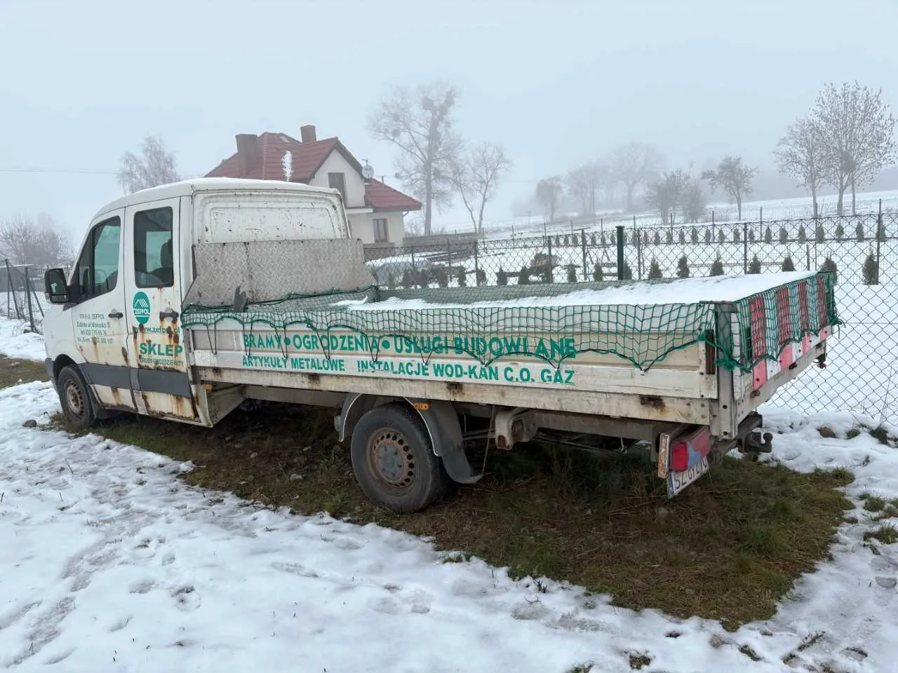 Biały samochód dostawczy z napisami na boku i tylnym zderzaku. Na boku drzwi widnieje napis 'SKLEP' oraz logo. Na boku platformy: 'BRAMY • OGRODZENIA • USŁUGI BUDOWLANE' oraz 'ARTYKUŁY METALOWE INSTALACJE WOD-KAN C.O. GAZ'. Platforma przykryta zieloną siatką, na niej leży śnieg. Samochód stoi na zasypanej śniegiem trawie, w tle dom z czerwonym dachem, ogrodzenie siatkowe, drzewa i mglisty krajobraz. Na tylnej tablicy rejestracyjnej widoczny jest fragment numeru '5207'.