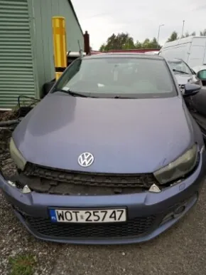 Niebieski samochód marki Volkswagen z przodu, z widocznym logo na masce i tablicą rejestracyjną WOT-25747. Pojazd stoi na żwirowym podłożu. W tle zielony budynek przemysłowy, żółta konstrukcja oraz inne pojazdy, w tym biały van. Niebo jest zachmurzone.