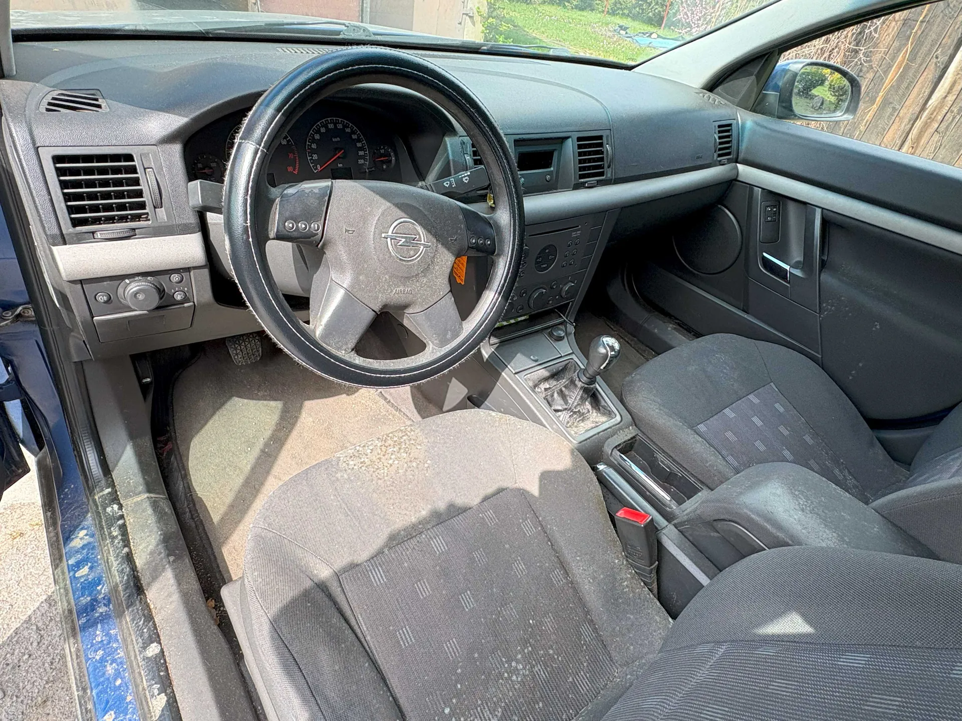 Wnętrze samochodu z kierownicą marki Opel z napisem "AIRBAG", tablicą rozdzielczą z licznikami prędkości i obrotomierzem, dźwignią zmiany biegów, przednimi siedzeniami w szarym kolorze z wzorem, otwartą drzwiami kierowcy o niebieskiej farbie oraz wentylacjami i przyciskami na desce rozdzielczej. Za przednią szybą widoczna trawa i ogrodzenie.