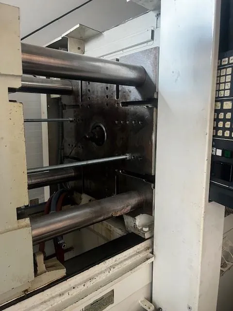 Przemysłowa maszyna z metalowymi cylindrami, białą konstrukcją i widocznymi śladami zużycia. Po prawej stronie panel sterowania z przyciskami i małym ekranem. W tle elementy instalacji technicznej.