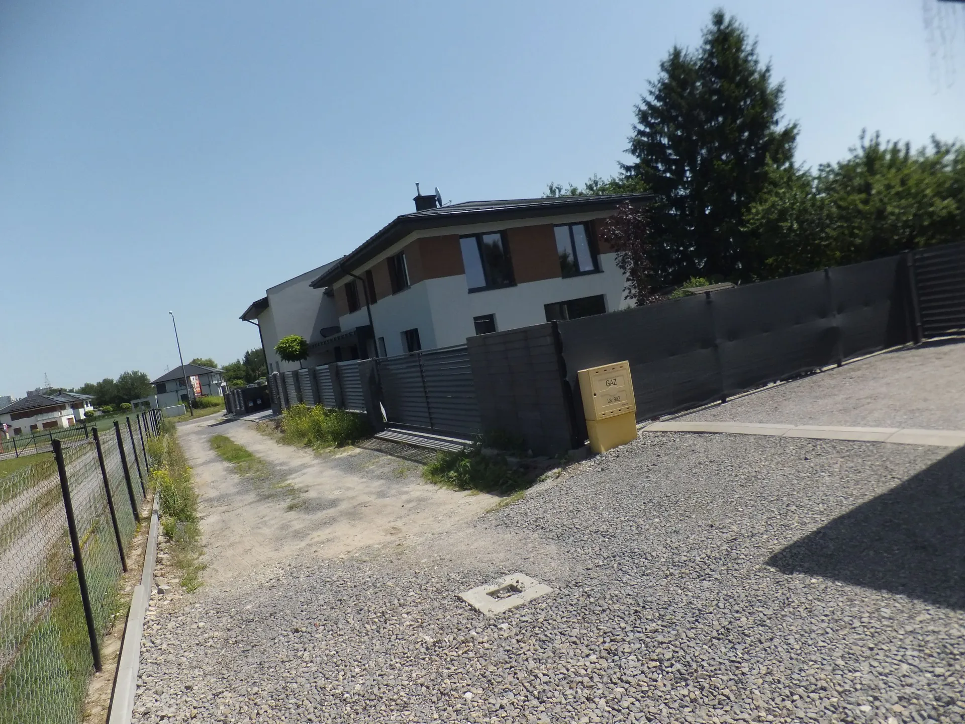 Widok na nowoczesny, biały dom jednorodzinny z ciemnym dachem i oknami, otoczony ciemnym, poziomym ogrodzeniem. Przed domem znajduje się żwirowy podjazd i piaszczysta droga. Po prawej stronie ogrodzenia stoi żółta skrzynka z napisem 'GAZ NR 802'. Po lewej stronie widoczny jest metalowy płot z siatki oraz fragmenty innych budynków w oddali pod bezchmurnym, niebieskim niebem.
