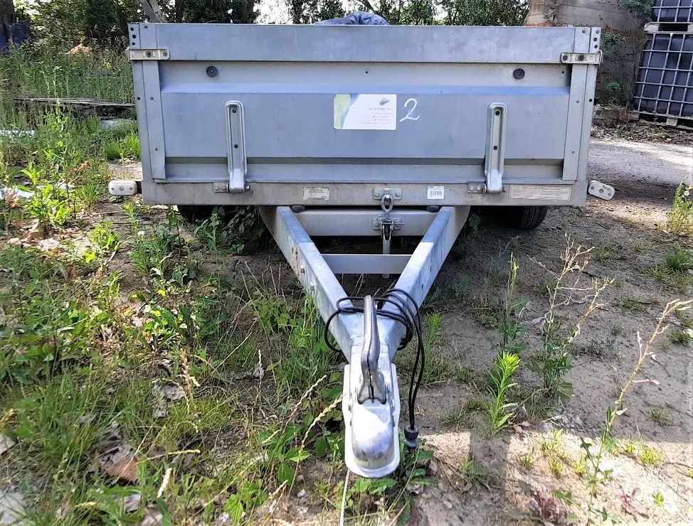 Srebrna, metalowa przyczepa transportowa z otwartą skrzynią, zaparkowana na trawiastym i piaszczystym podłożu. Na tylnej ścianie przyczepy znajduje się biała naklejka z cyfrą 2. Widoczny jest dyszel z zaczepem oraz kable elektryczne.