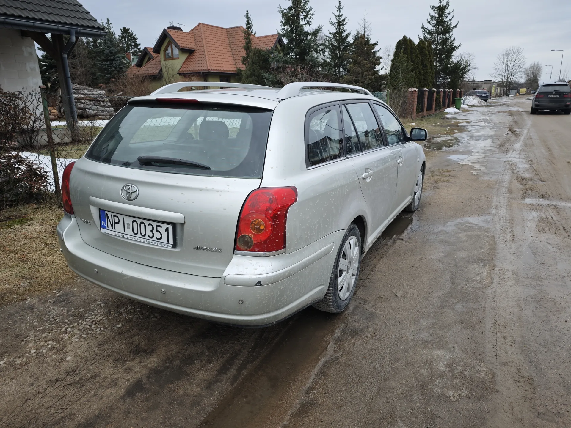Srebrny samochód osobowy typu kombi marki Toyota Avensis zaparkowany na błotnistej drodze. Widoczny tył pojazdu z tablicą rejestracyjną PL NP 00351. Samochód ma dachowy bagażnik i czerwone światła tylnego. W tle domy z czerwonymi dachami, drzewa, ogrodzenie oraz drugi samochód na drodze. Droga pokryta jest kałużami i resztkami śniegu, niebo jest zachmurzone.