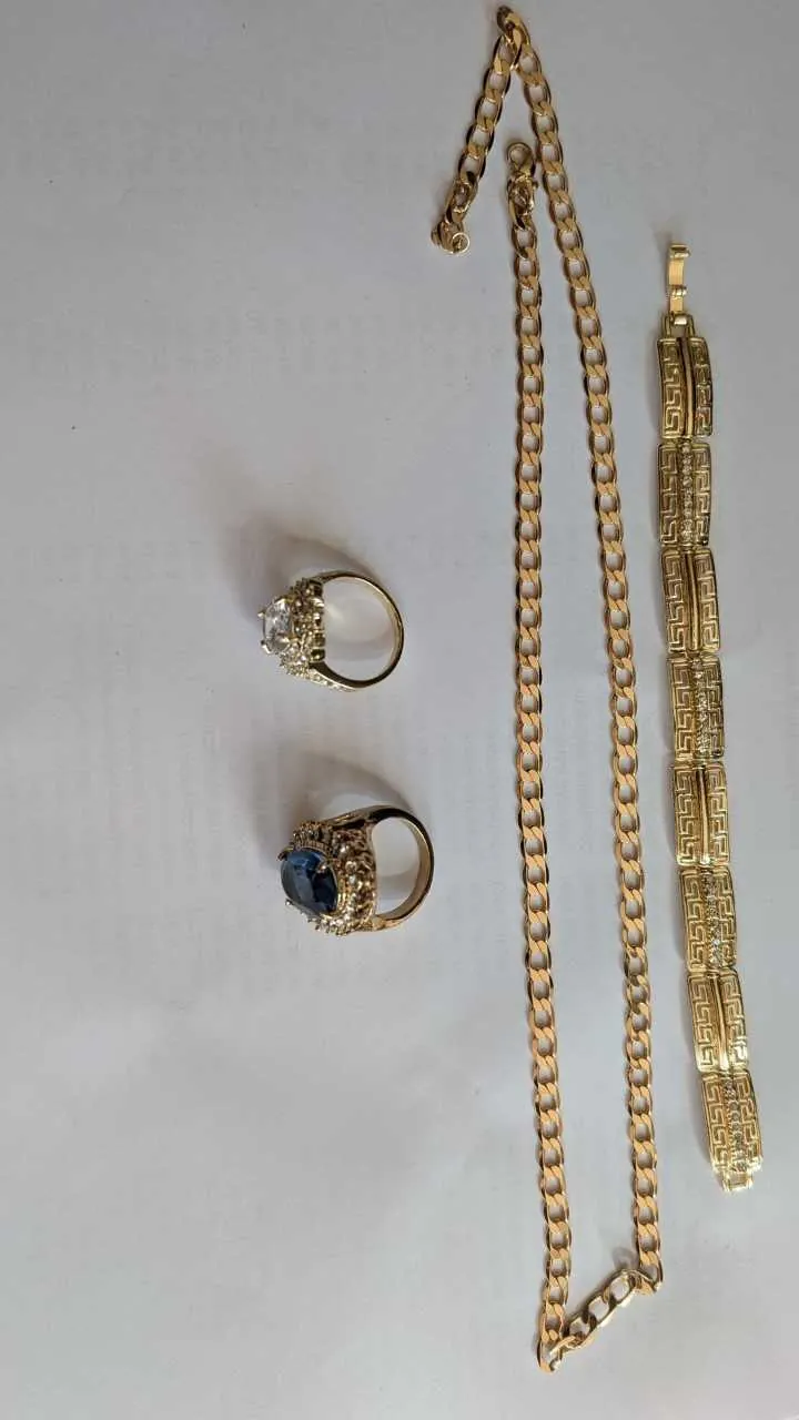 Na białym tle umieszczono cztery elementy biżuterii: dwa pierścionki, łańcuszek i bransoletkę. Pierścionki mają złote obrączki; górny z brylantem, dolny z niebieskim kamieniem otoczonym drobnymi kryształkami. Długi łańcuszek z grubymi ogniwnami znajduje się po prawej stronie pierścionków. Obok niego leży bransoletka z geometrycznym wzorem i drobnymi kryształkami. Wszystkie elementy są ułożone pionowo.