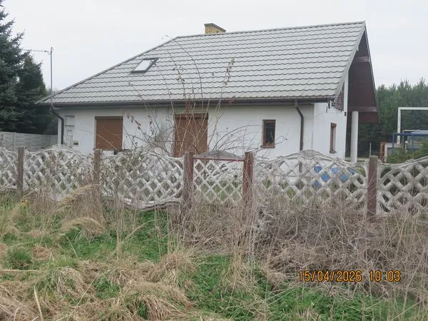Biały, parterowy dom z szarym, spadzistym dachem i jednym oknem dachowym. Na fasadzie widoczne są dwa brązowe rolety w oknach oraz mniejsze okno po prawej stronie. Przed domem znajduje się biały, ażurowy płot z betonowymi słupkami, za którym rosną wysokie, suche trawy i krzewy. W prawym dolnym rogu zdjęcia widnieje data i godzina: 15/04/2026 10:03.