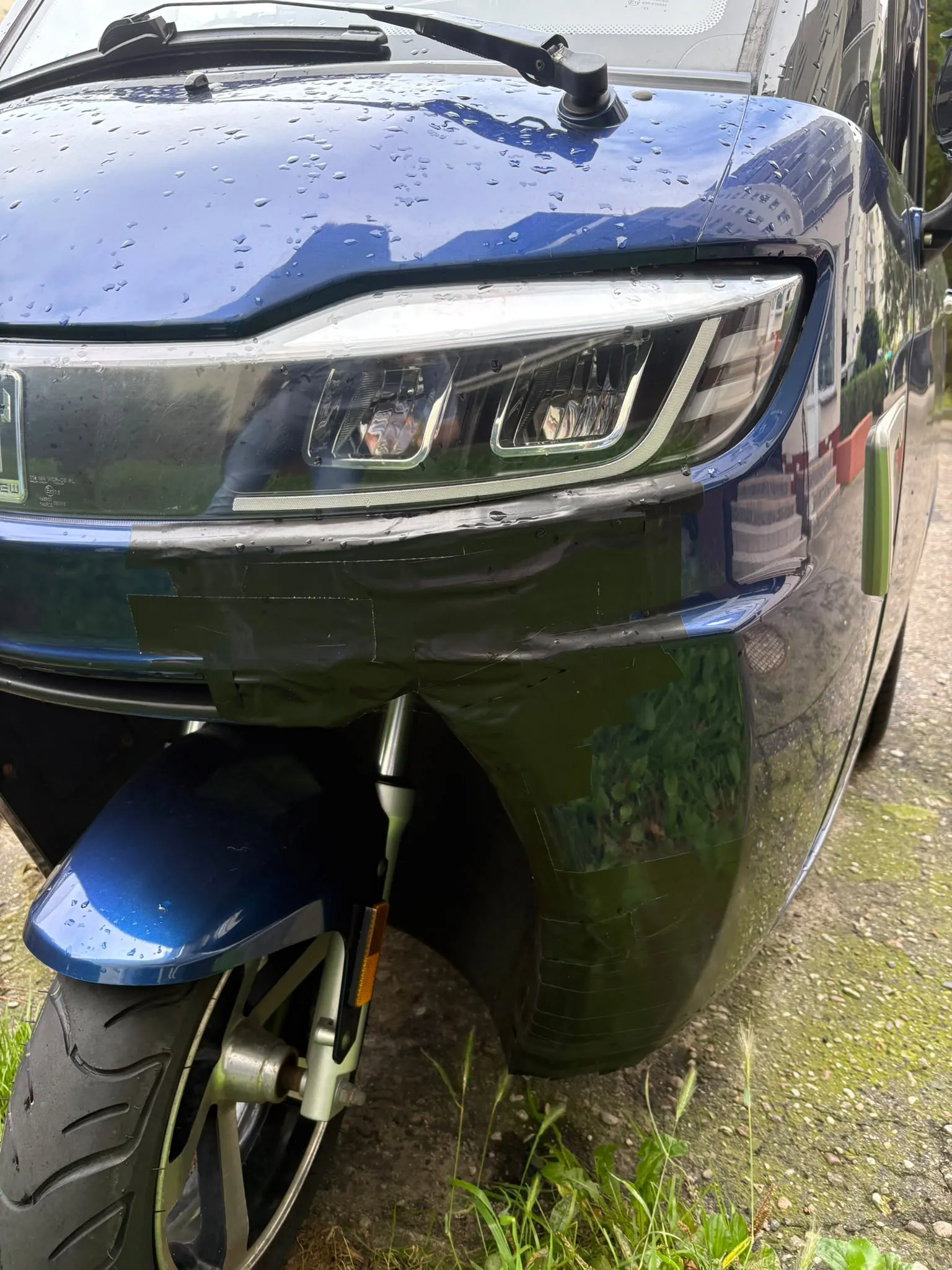 Zbliżenie na przód niebieskiego skutera. Widoczny jest reflektor, nad którym znajduje się mokra obudowa z kroplami wody. Poniżej reflektora plastikowy element karoserii jest uszkodzony i prowizorycznie sklejony szeroką, szarą taśmą klejącą. W dolnej części zdjęcia widoczne jest przednie koło z oponą oraz błotnik.