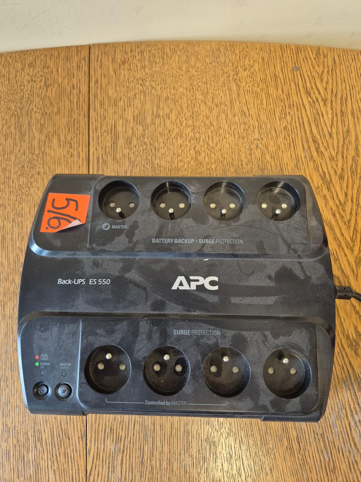 Czarny przedłużacz z listwą zasilającą marki APC, model Back-UPS ES 550. Urządzenie posiada osiem gniazd elektrycznych typu E (po cztery w dwóch rzędach). Na górnej części znajduje się napis 'BATTERY BACKUP + SURGE PROTECTION', a na dolnej 'SURGE PROTECTION'. Po lewej stronie widoczne są diody kontrolne 'POWER' oraz 'MASTER', a pod nimi przycisk. Na obudowie znajduje się pomarańczowa naklejka z napisem '516'. Urządzenie leży na drewnianym blacie.